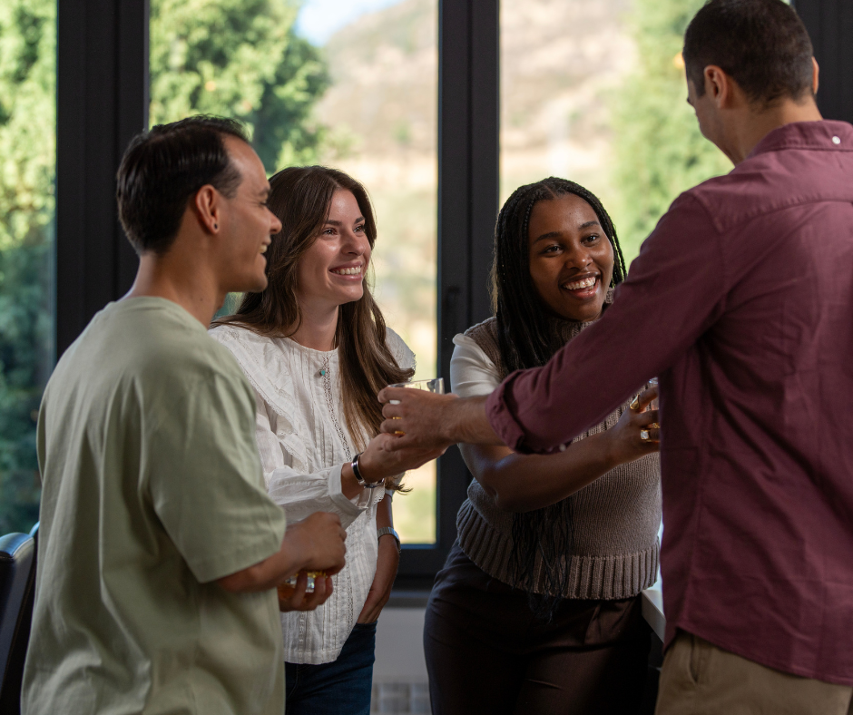 Four people socializing indoors with drinks, smiling, near a large window with a green outdoor background.