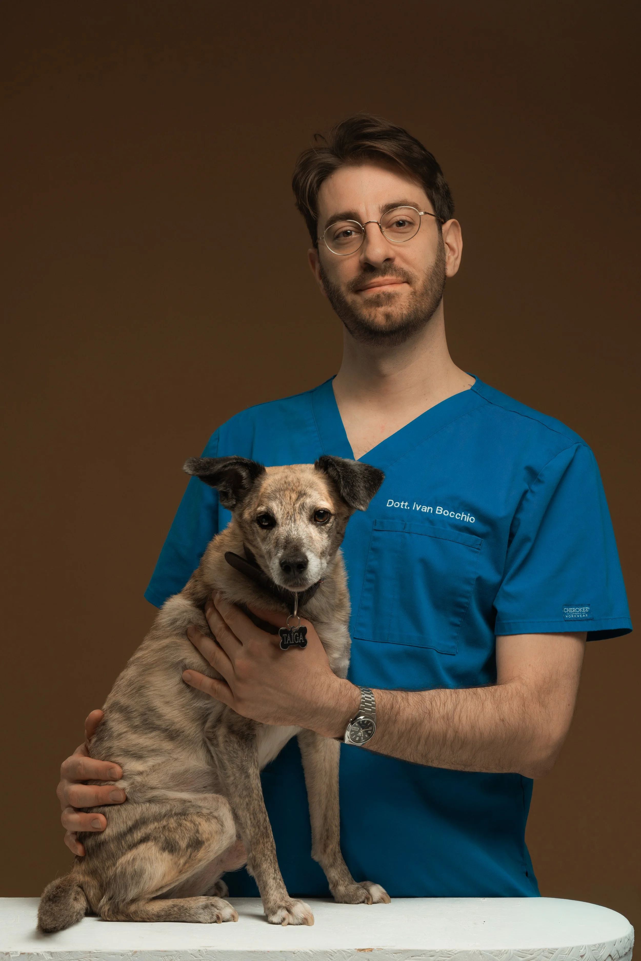 Veterinario con occhiali e camice blu che abbraccia un cane da compagnia su un tavolo.