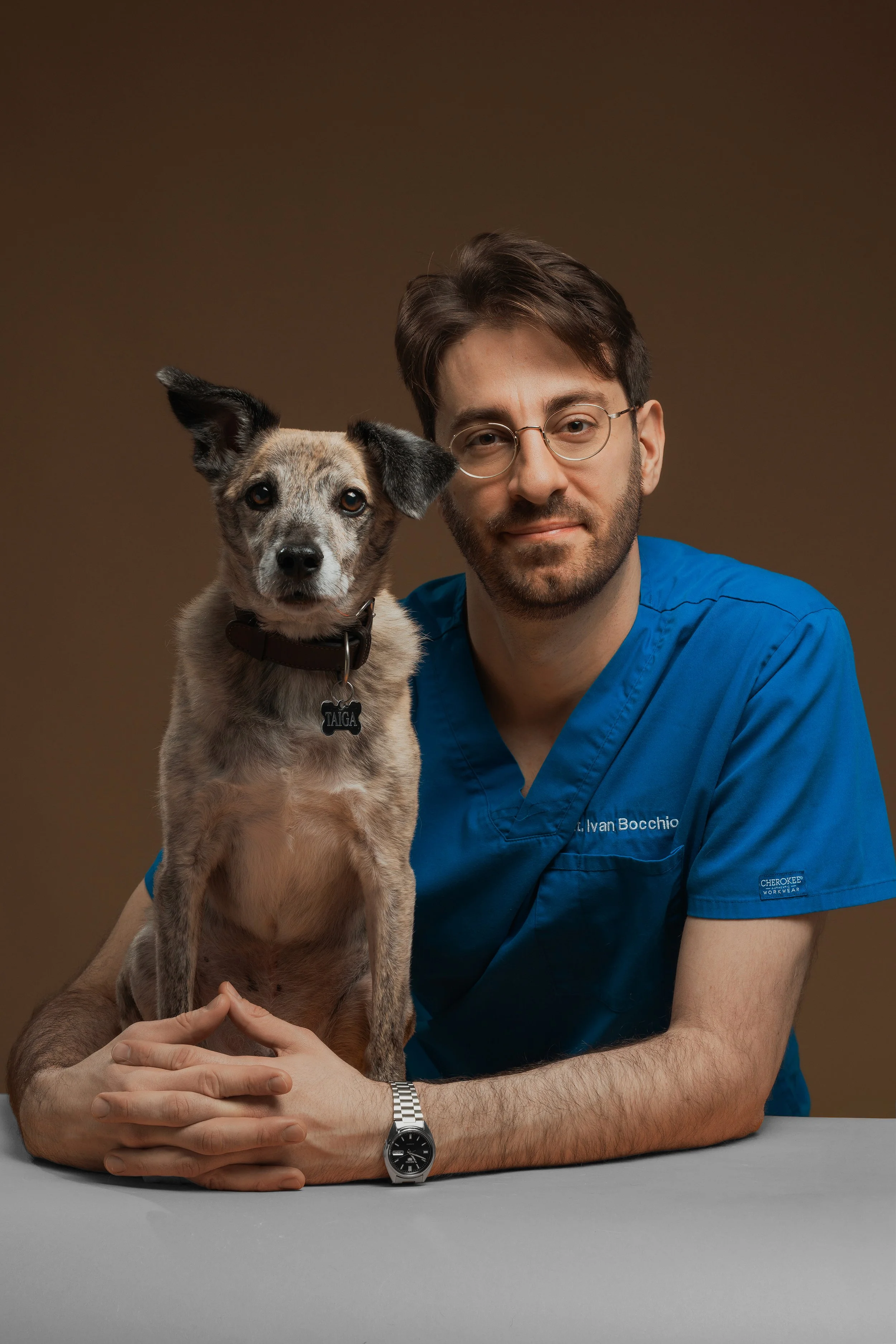 Veterinario in camice blu con cane a pelo corto e maculato, seduti davanti a uno sfondo marrone.