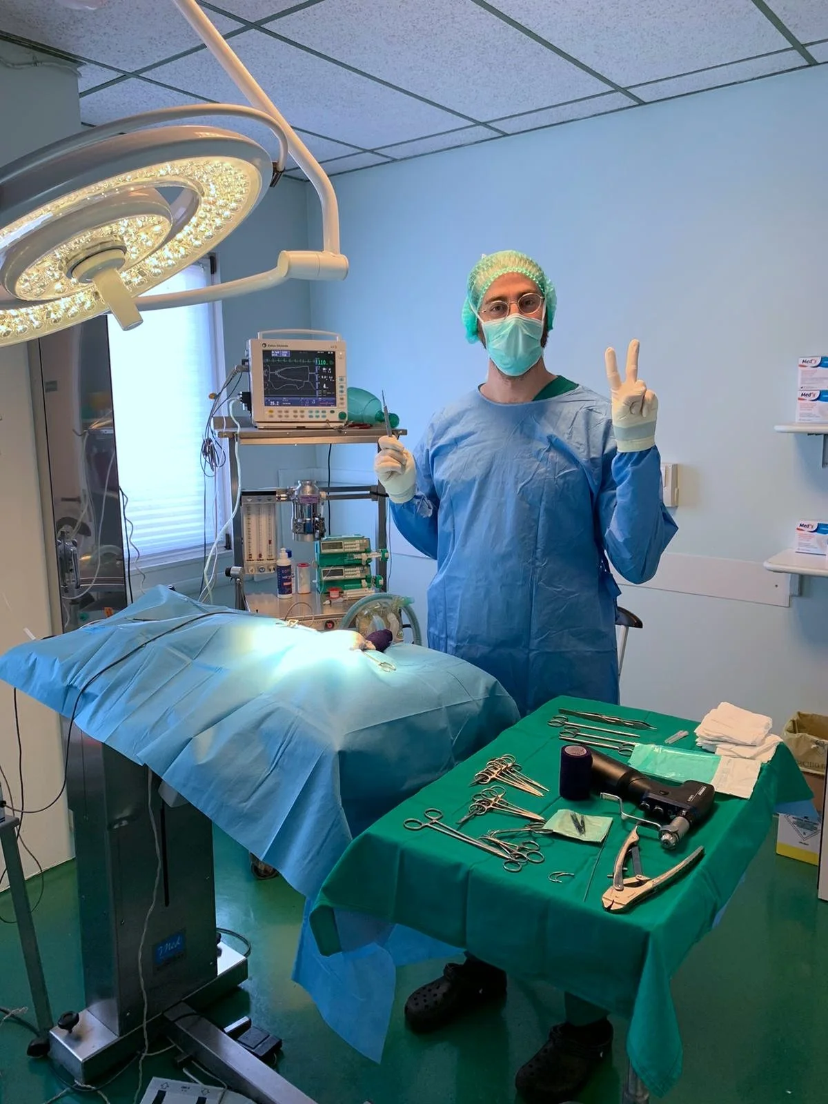 Medico veterinario in sala operatoria che fa un gesto di pace con le dita, con strumenti chirurgici e apparecchiature mediche sul tavolo e sullo sfondo.