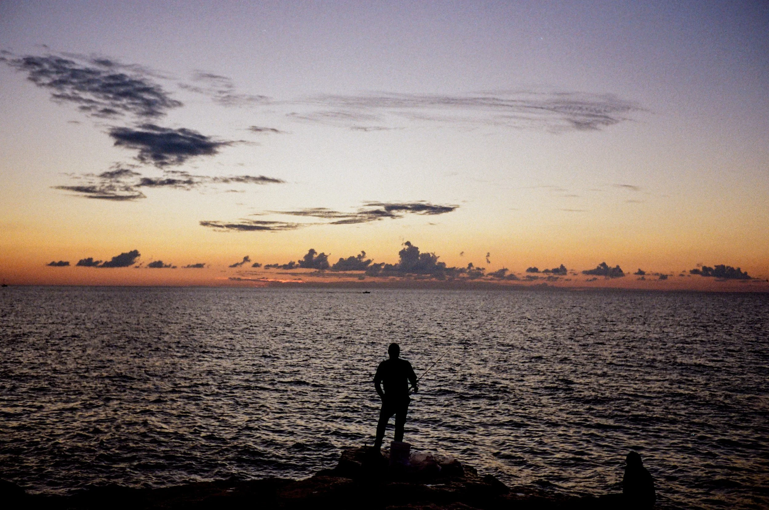 Silhouette of a person fishing on rocks by the ocean at sunset with a cloudy sky and colorful horizon.