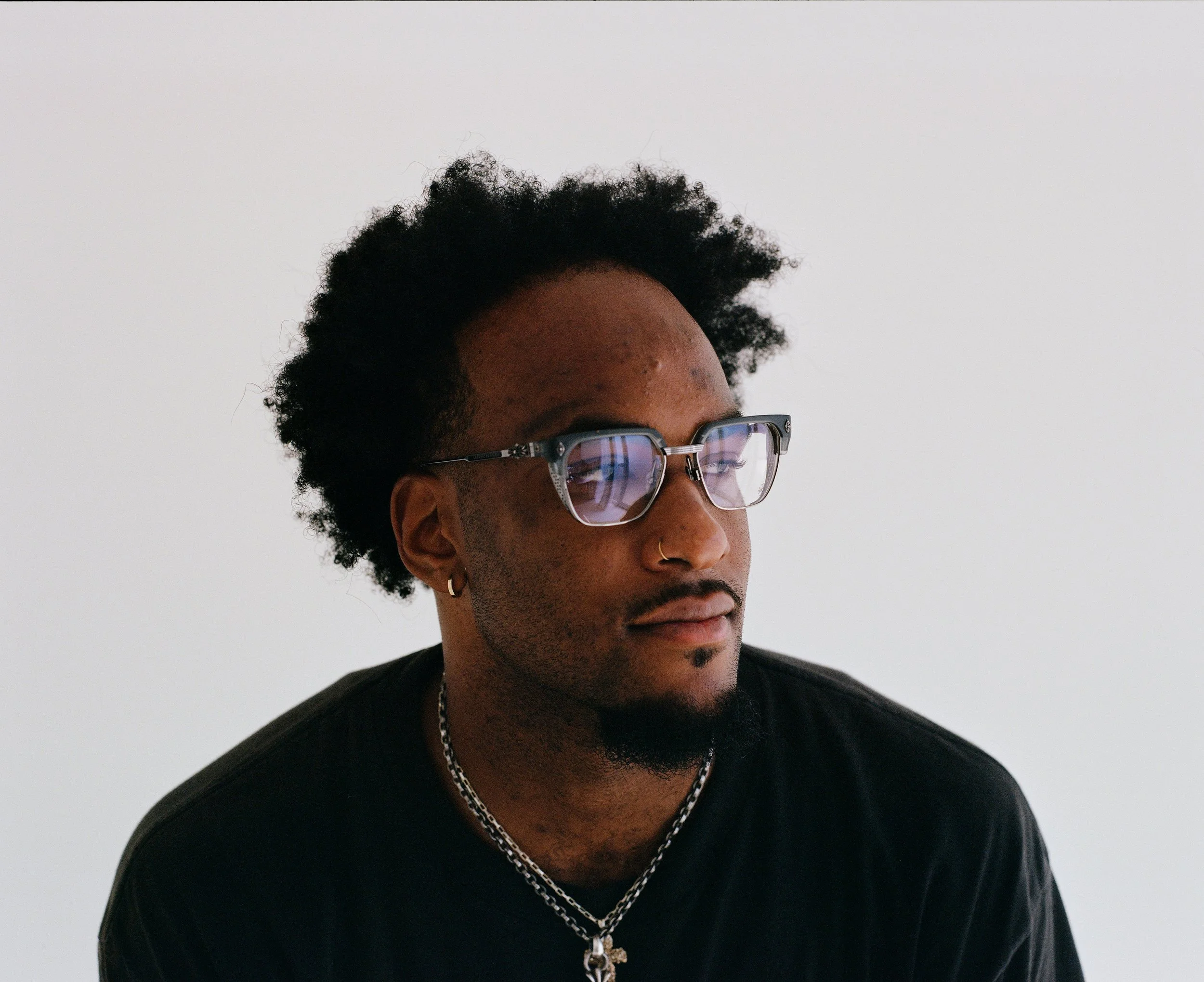 A young man with dark skin, curly hair, and glasses, wearing a nose ring and earrings, looking to the side against a plain white background.