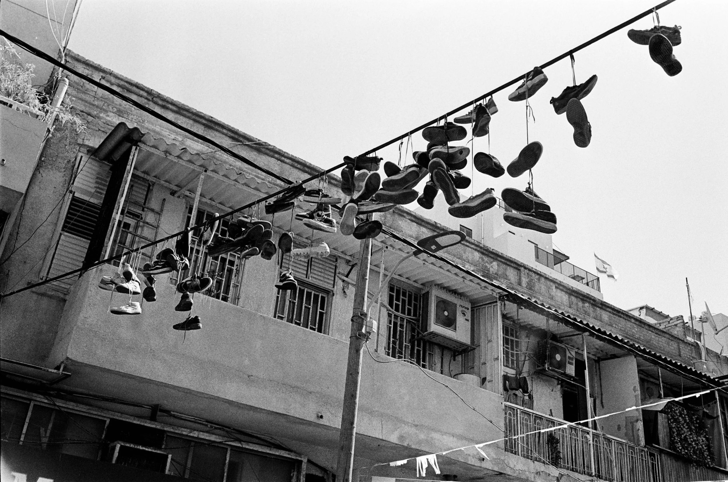 Multiple pairs of shoes hanging on clotheslines outside, in front of an apartment building with balconies and air conditioning units, black and white photo.