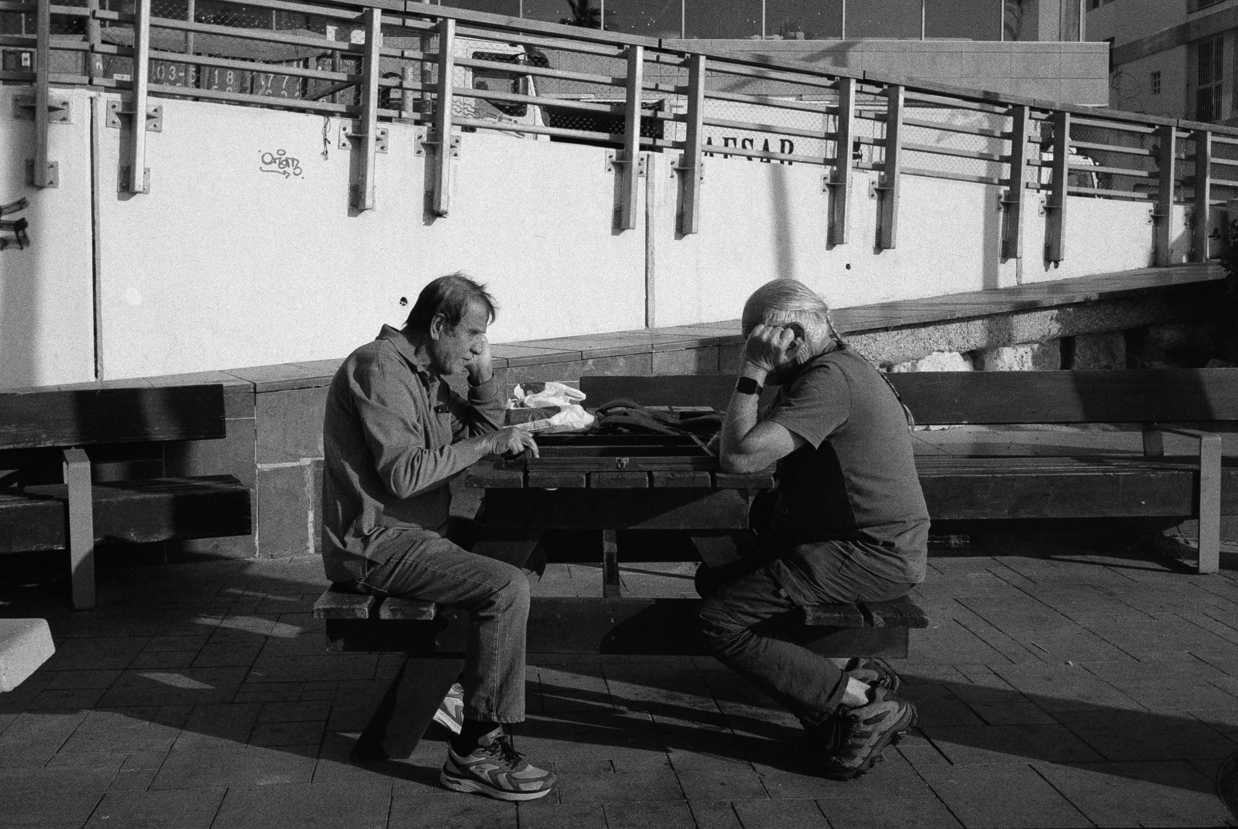 Two men sitting at an outdoor picnic table, both with their heads resting on their hands, engaged in a serious or contemplative moment.