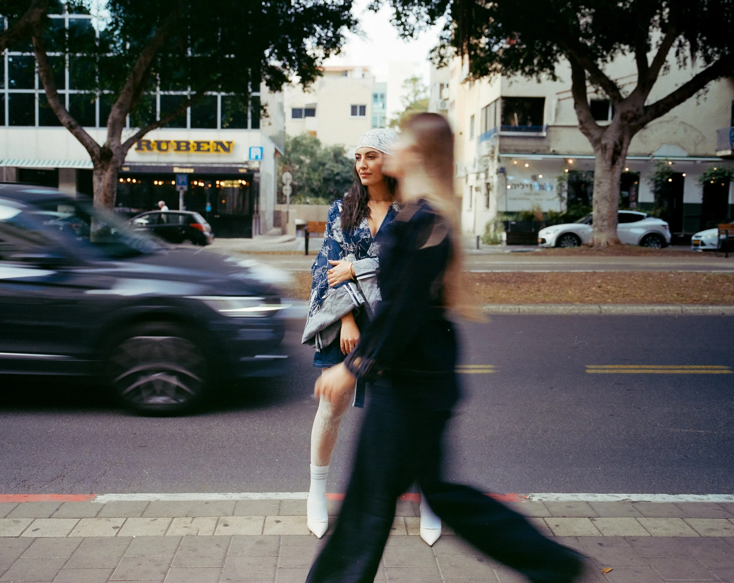 A woman with long dark hair, wearing a patterned blue dress, white hat, and white high-heeled boots, stands on the sidewalk in front of a blurred pedestrian and moving car, with trees and buildings in the background.