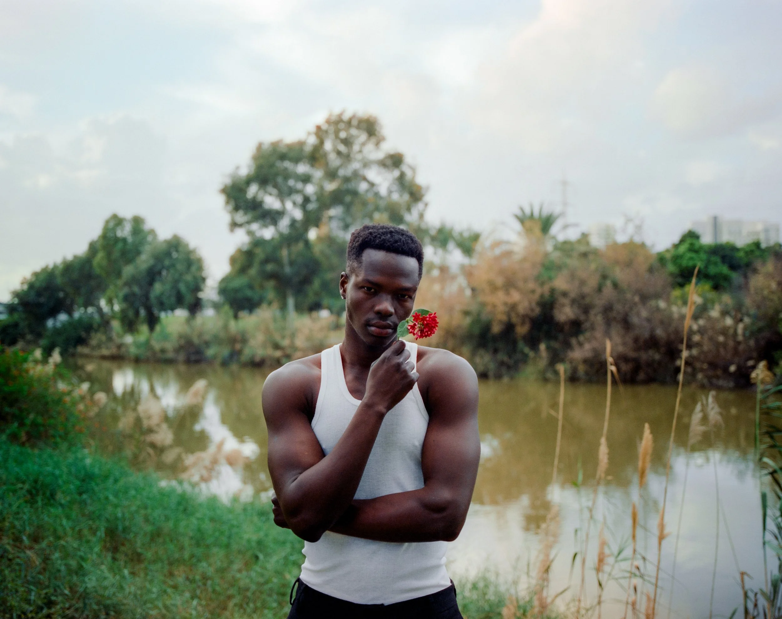 A young man with dark skin and short hair standing outdoors by a river, holding a small red flower near his face, wearing a white sleeveless shirt.