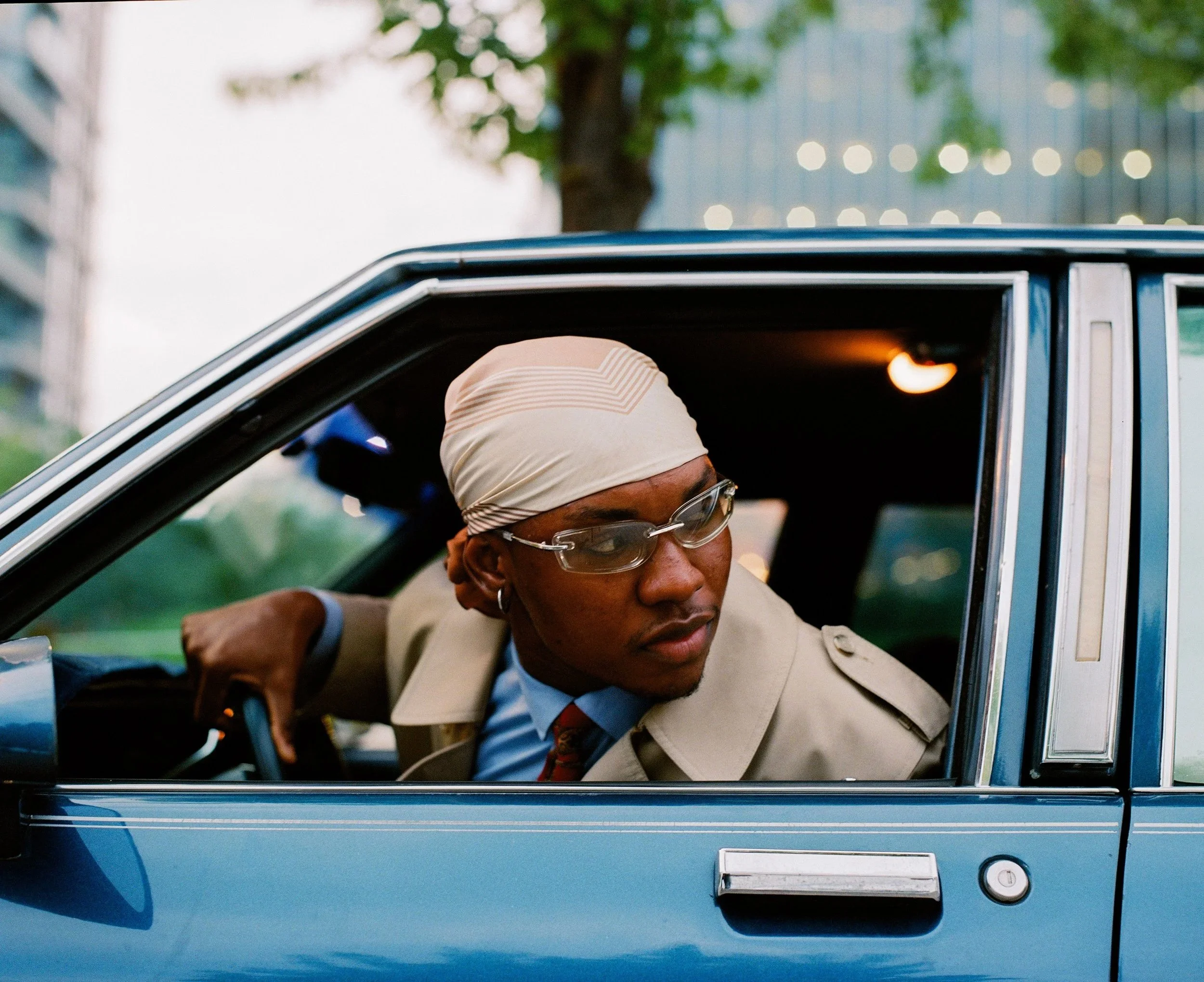 A man wearing glasses and a beige headscarf looks out from the driver's side of a vintage blue car, with a cityscape and trees in the background.