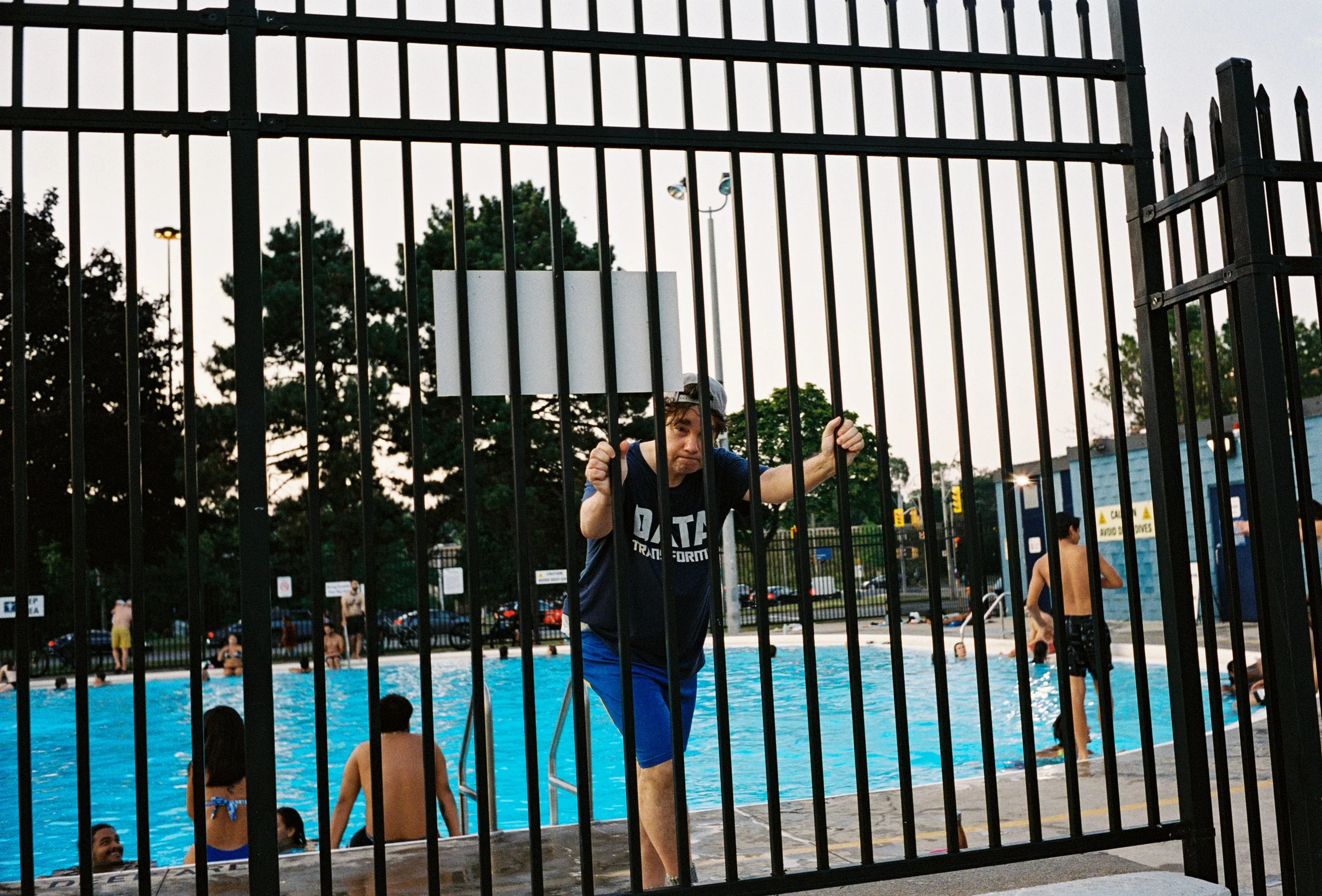 A man in blue shorts and a black T-shirt standing behind a black metal gate at a public swimming pool, holding onto the gate with both hands, with other people swimming and relaxing in the pool in the background.