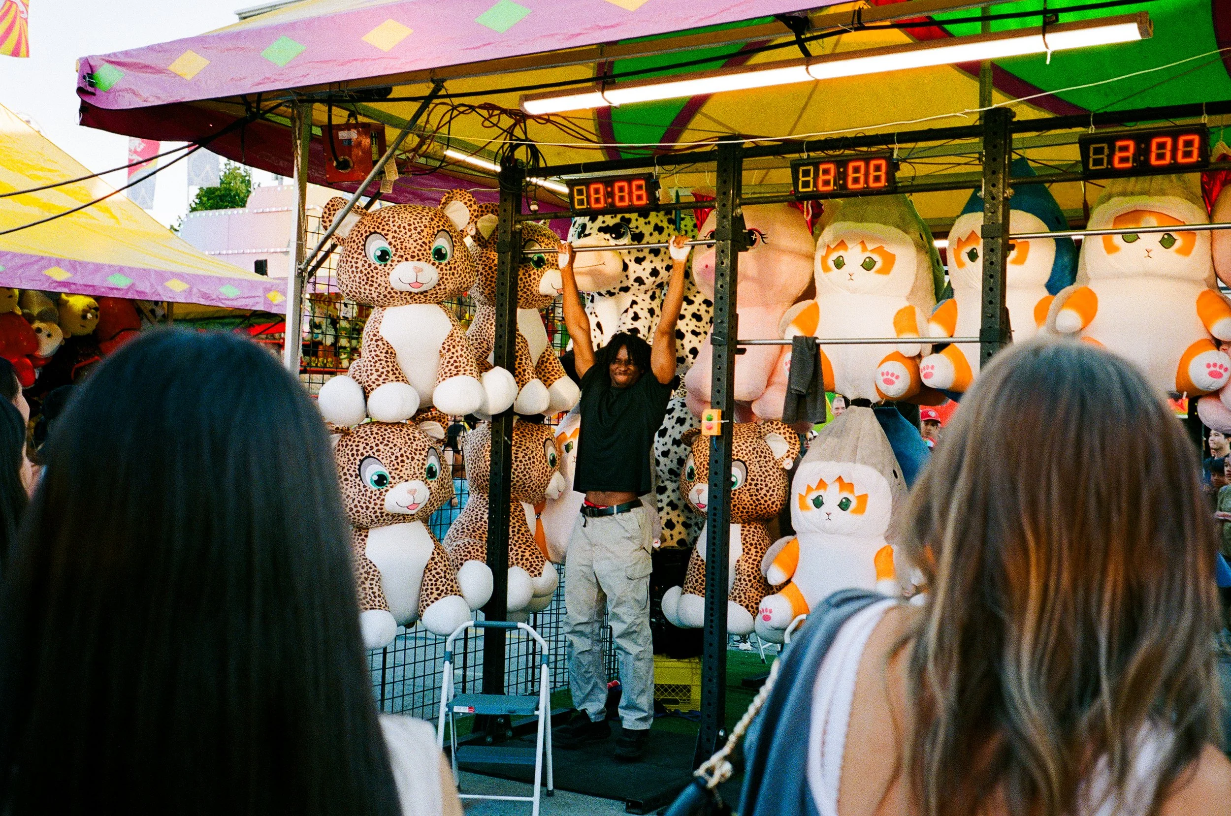 A carnival game booth with a man hanging from a bar, surrounded by plush animal toys, including leopards and cats, at an outdoor fair with people watching.