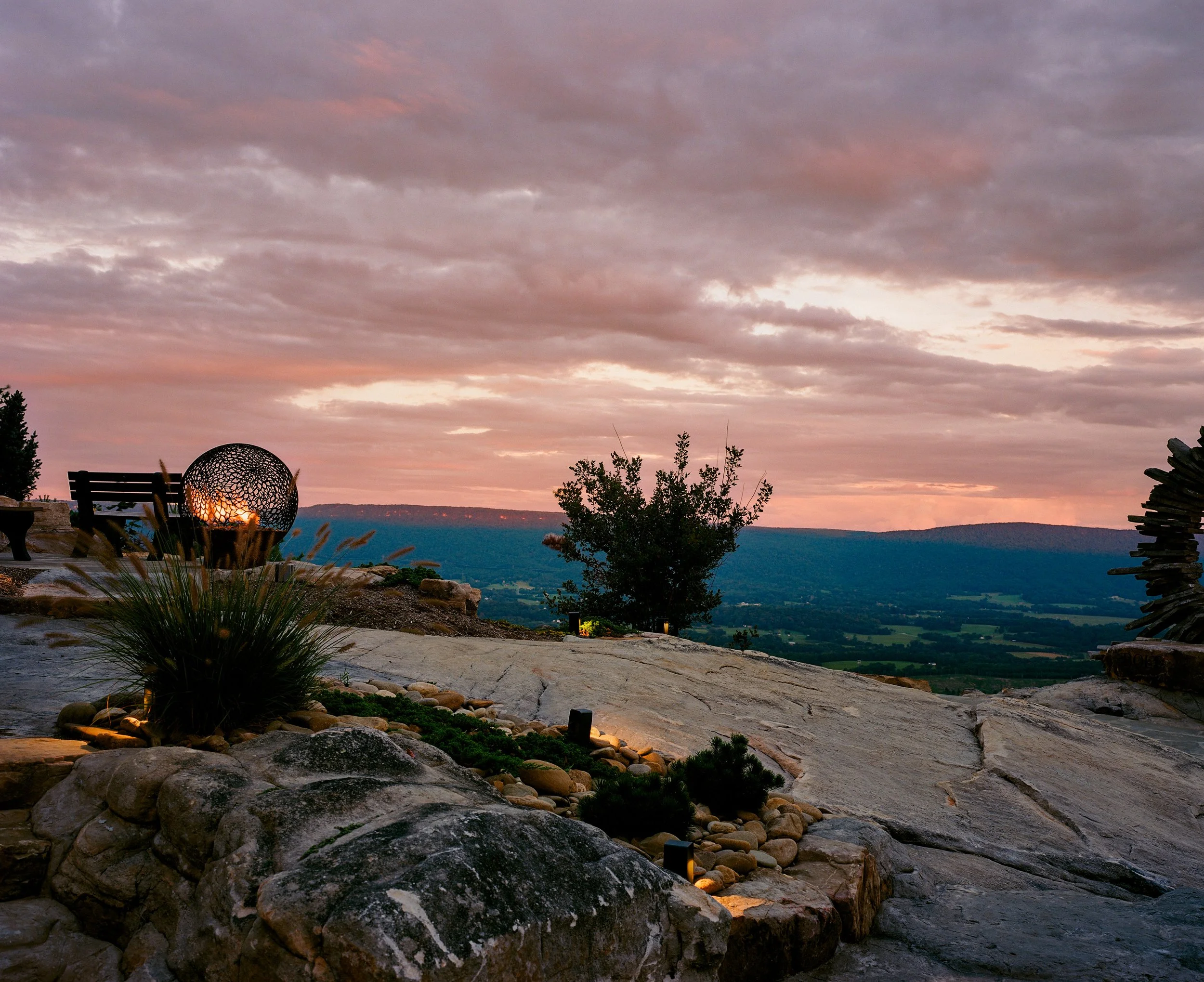 A scenic view of a sunset over a valley, with outdoor seating and decorative lighting on a rocky terrace.