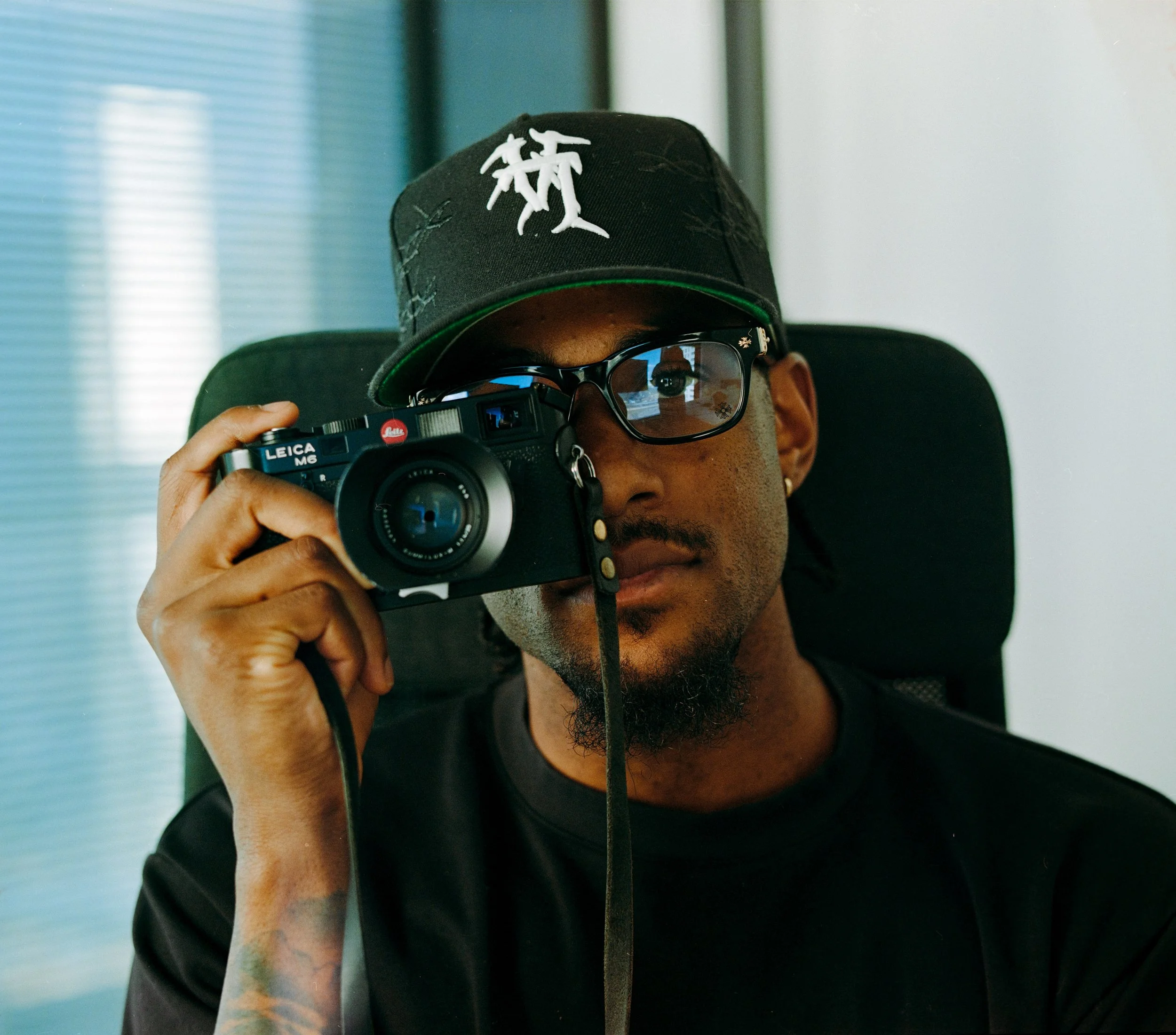 Man wearing black cap with white design, black glasses, holding a Leica camera with one eye, sitting in office chair in front of window.
