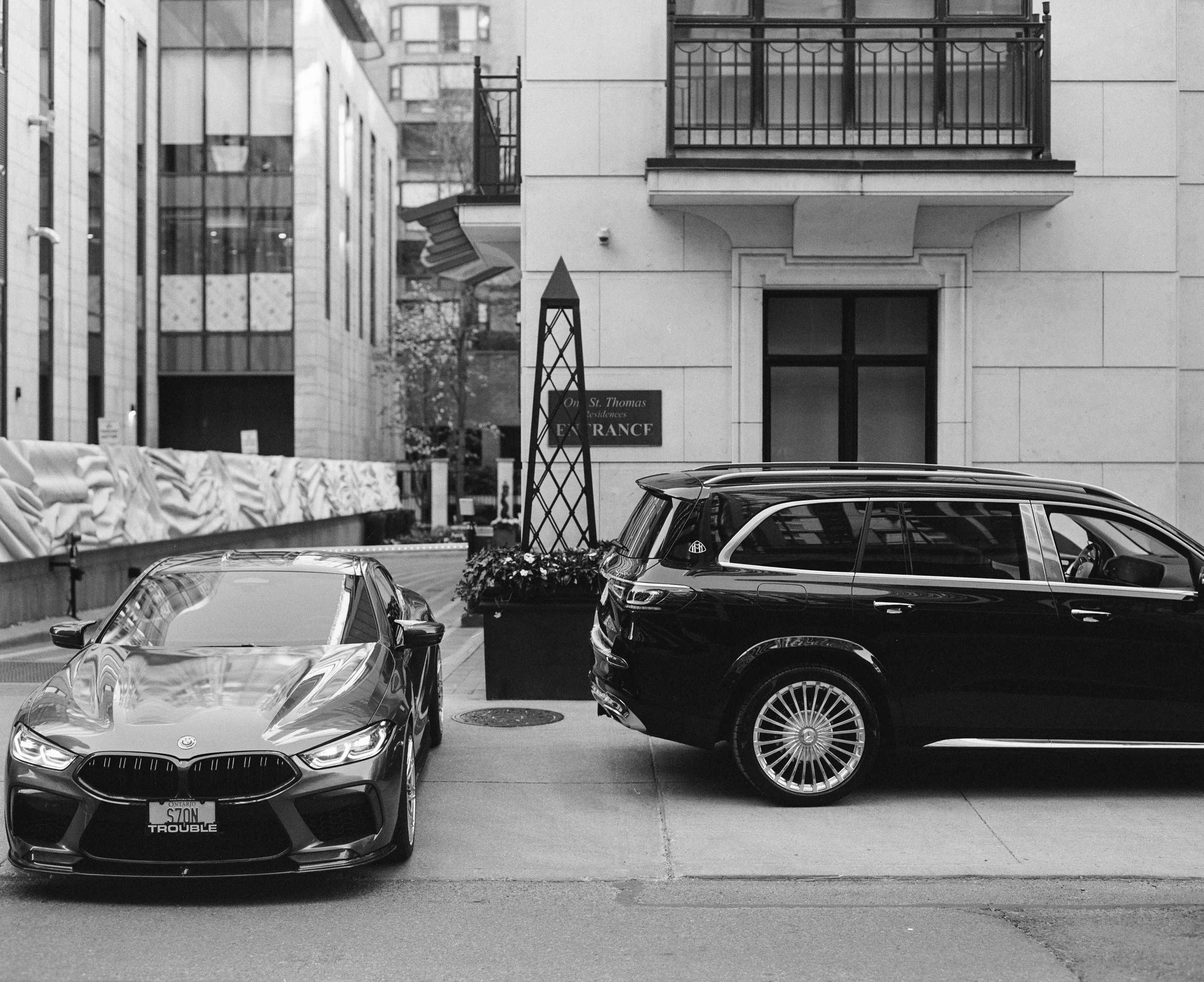 A black luxury SUV and a gray sports car parked on a city street in front of a beige building with balconies and a sign that reads "St. Thomas Residences Entrance."