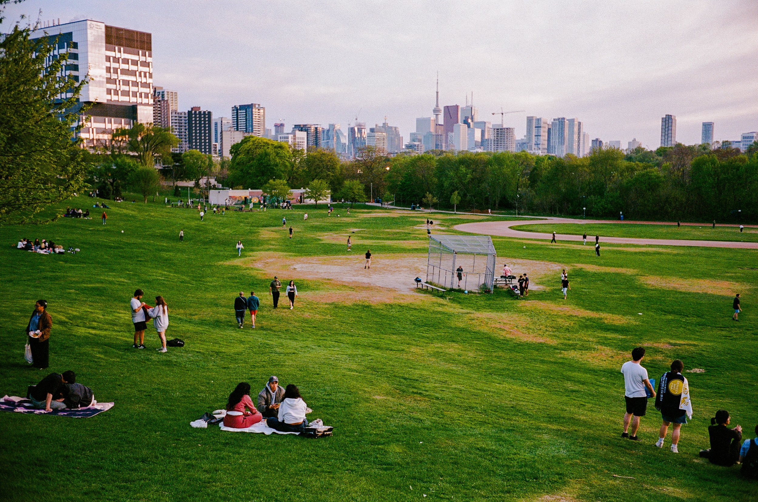 People enjoying a park with green grass, trees, and a city skyline in the background, some sitting on blankets, others walking or playing.