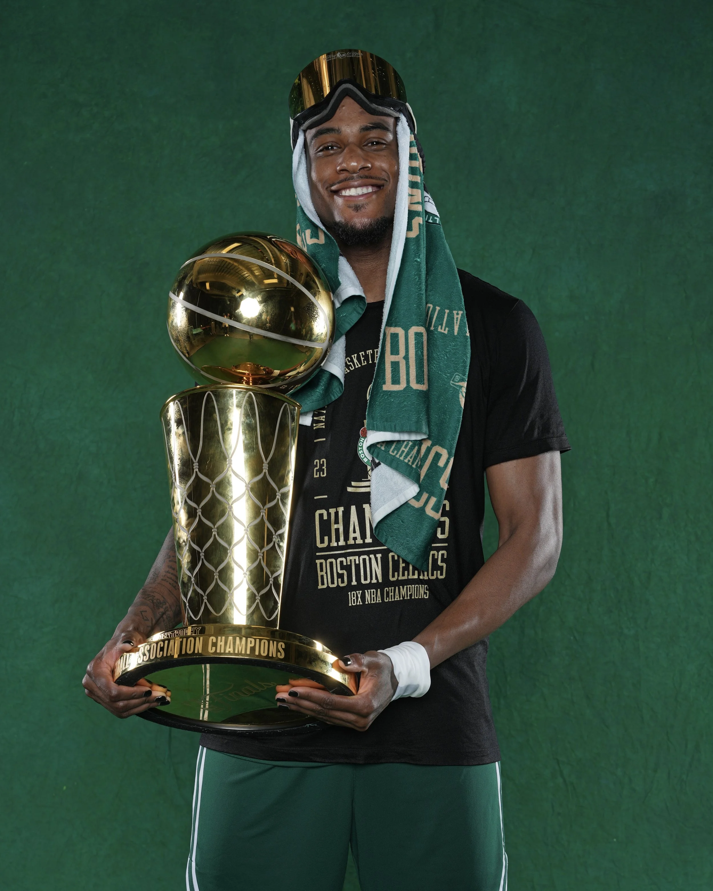 A male basketball player in a black t-shirt and green shorts holding a gold trophy, wearing a towel around his neck and a modified goggles with a golden lens, standing against a green background.