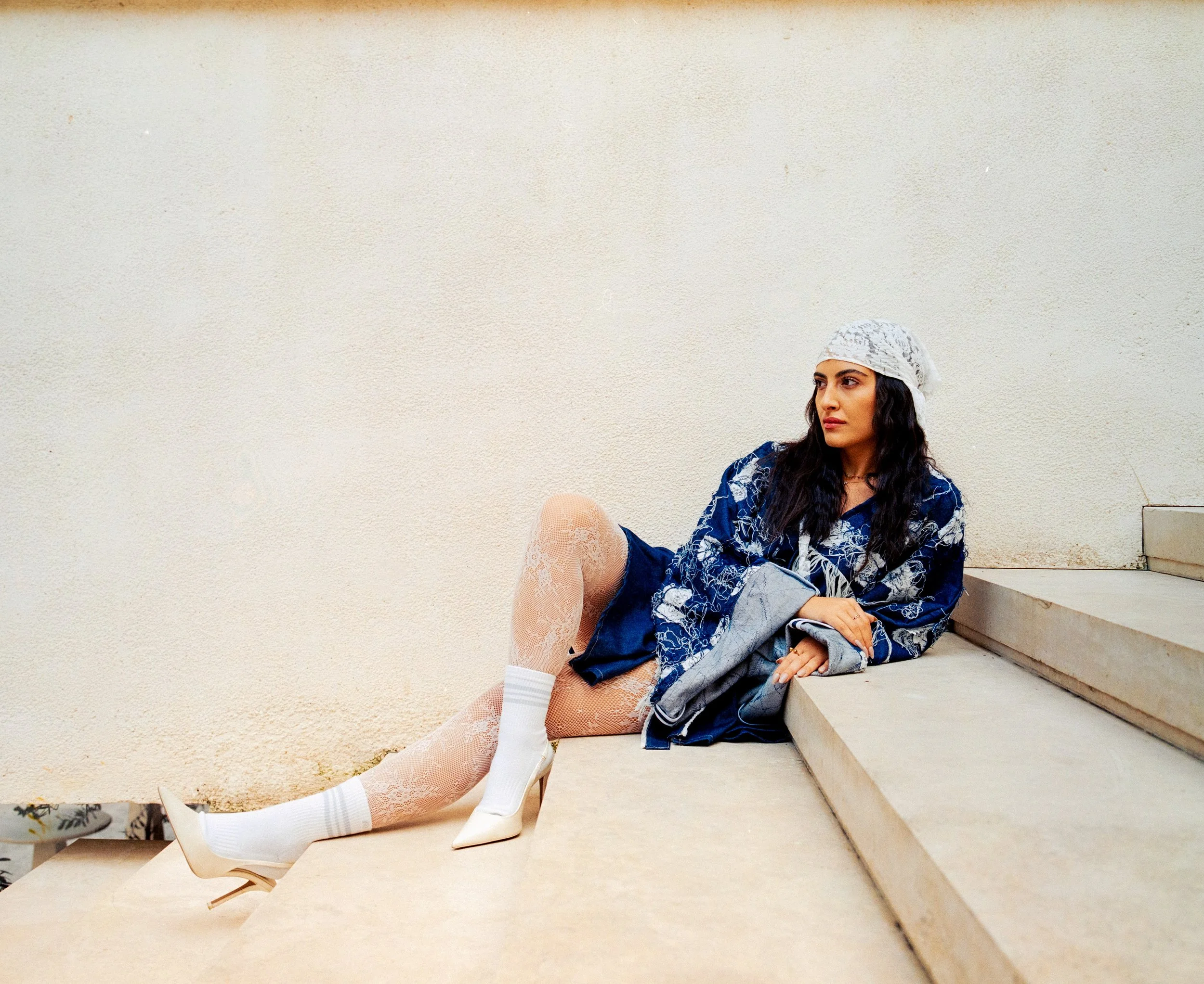 Woman with long dark hair wearing a white lace headband and denim jacket with floral patterns, sitting on outdoor steps with a neutral wall background.