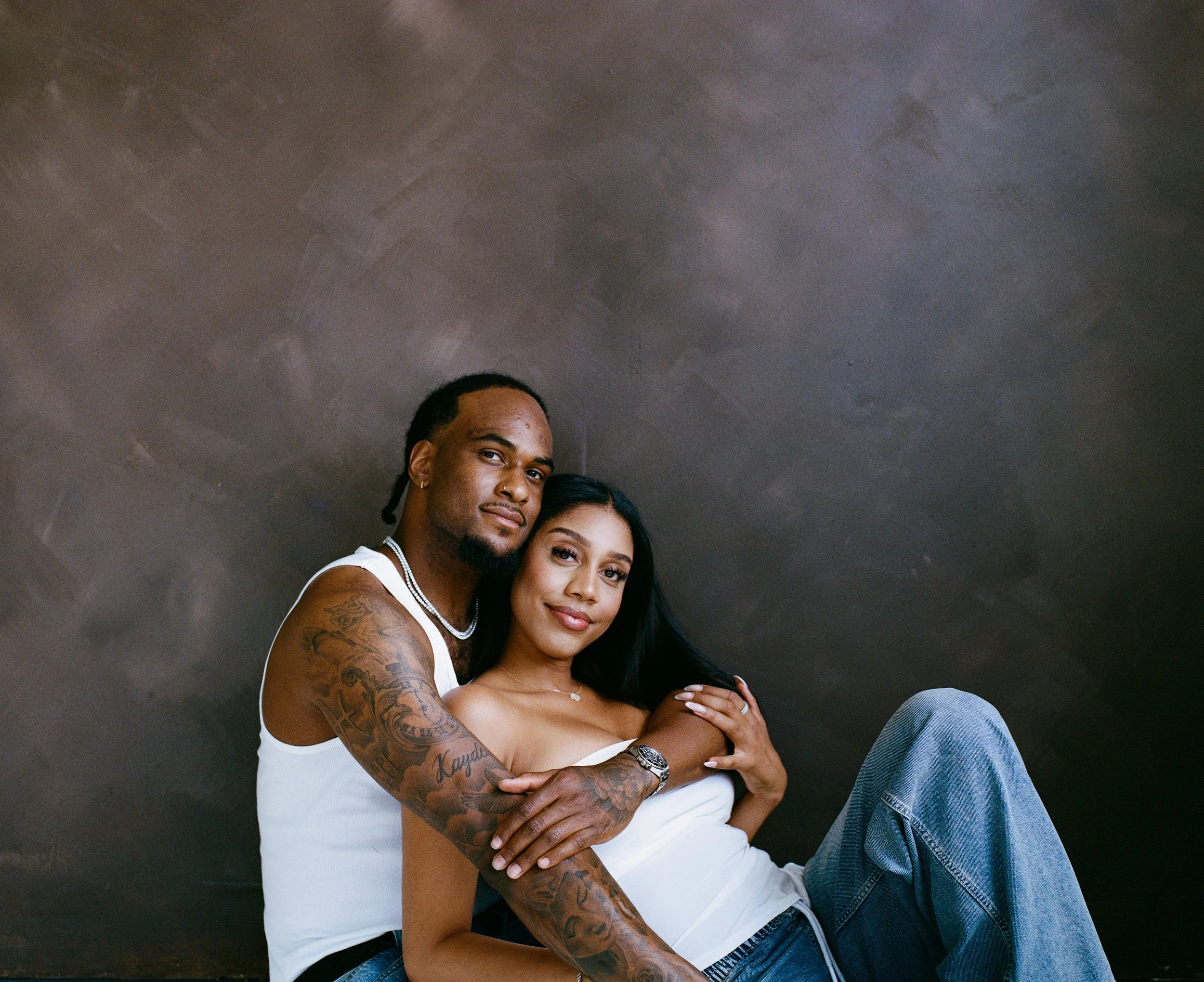 A young couple sitting close together and smiling, with a plain dark wall background. The man has tattoos on his right arm, wears a white tank top and jewelry. The woman has long black hair, wears a white strapless top and jeans.