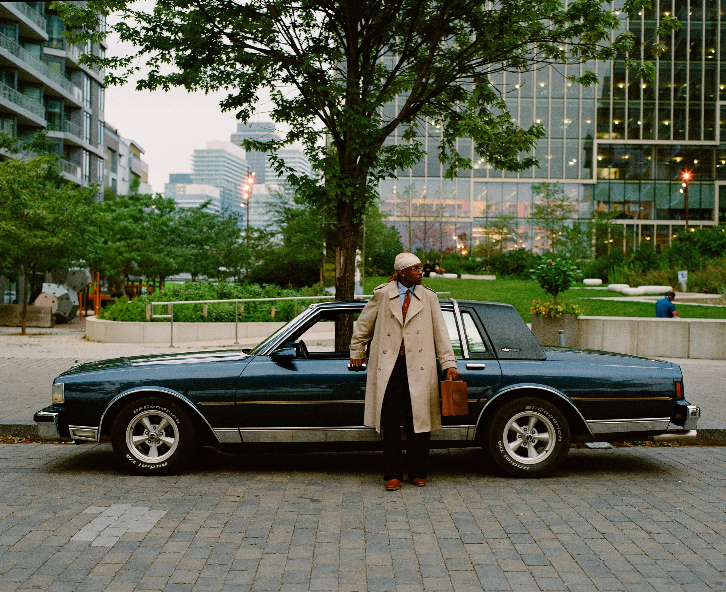 A man in a beige trench coat holding a brown briefcase stands next to a black vintage car in an urban park with modern buildings in the background.