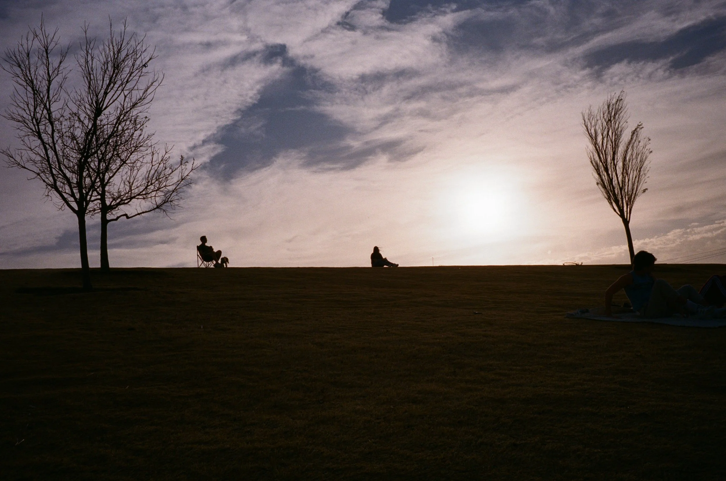 Silhouette of four people with backpacks sitting on a grassy hill at sunset, two on chairs and two lying on the ground, with bare trees and a cloudy sky in the background.
