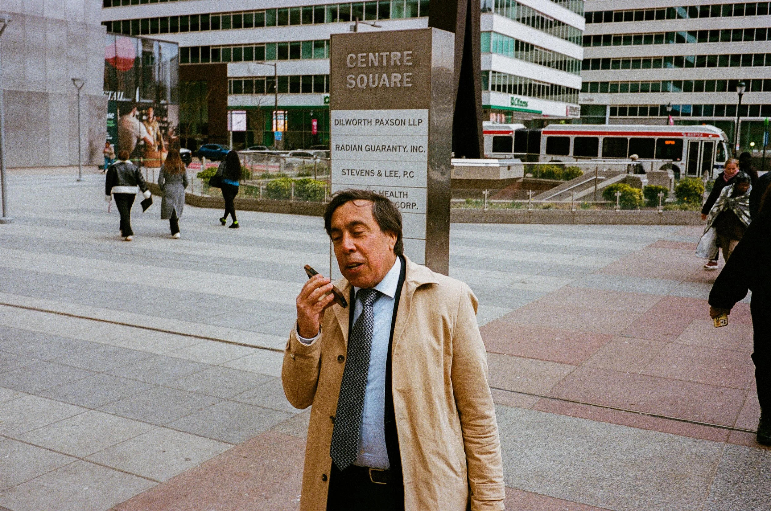 A man in a beige trench coat and tie talking on a cell phone in an urban plaza with a large sign marked 'Centre Square' behind him, with several people walking around and modern office buildings in the background.