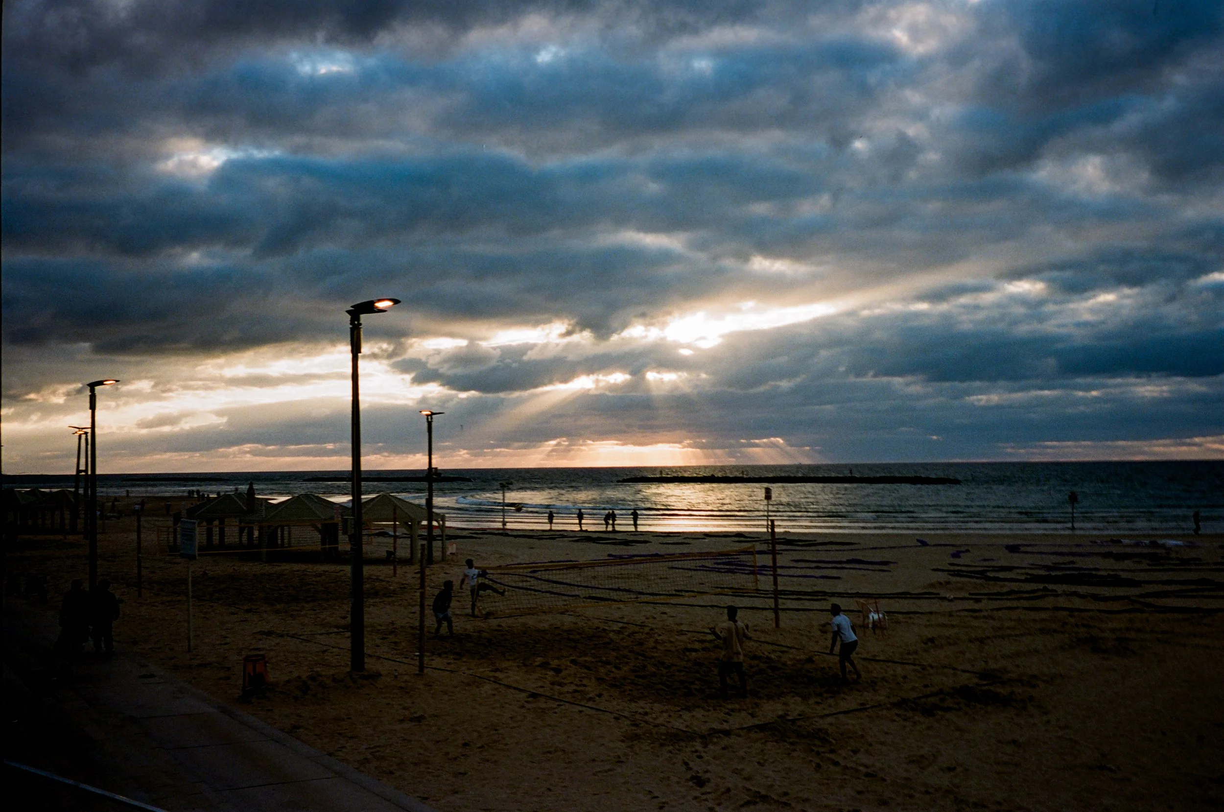 A beach scene at sunset or sunrise with a cloudy sky, several people playing volleyball on a sandy court, and the ocean in the background.