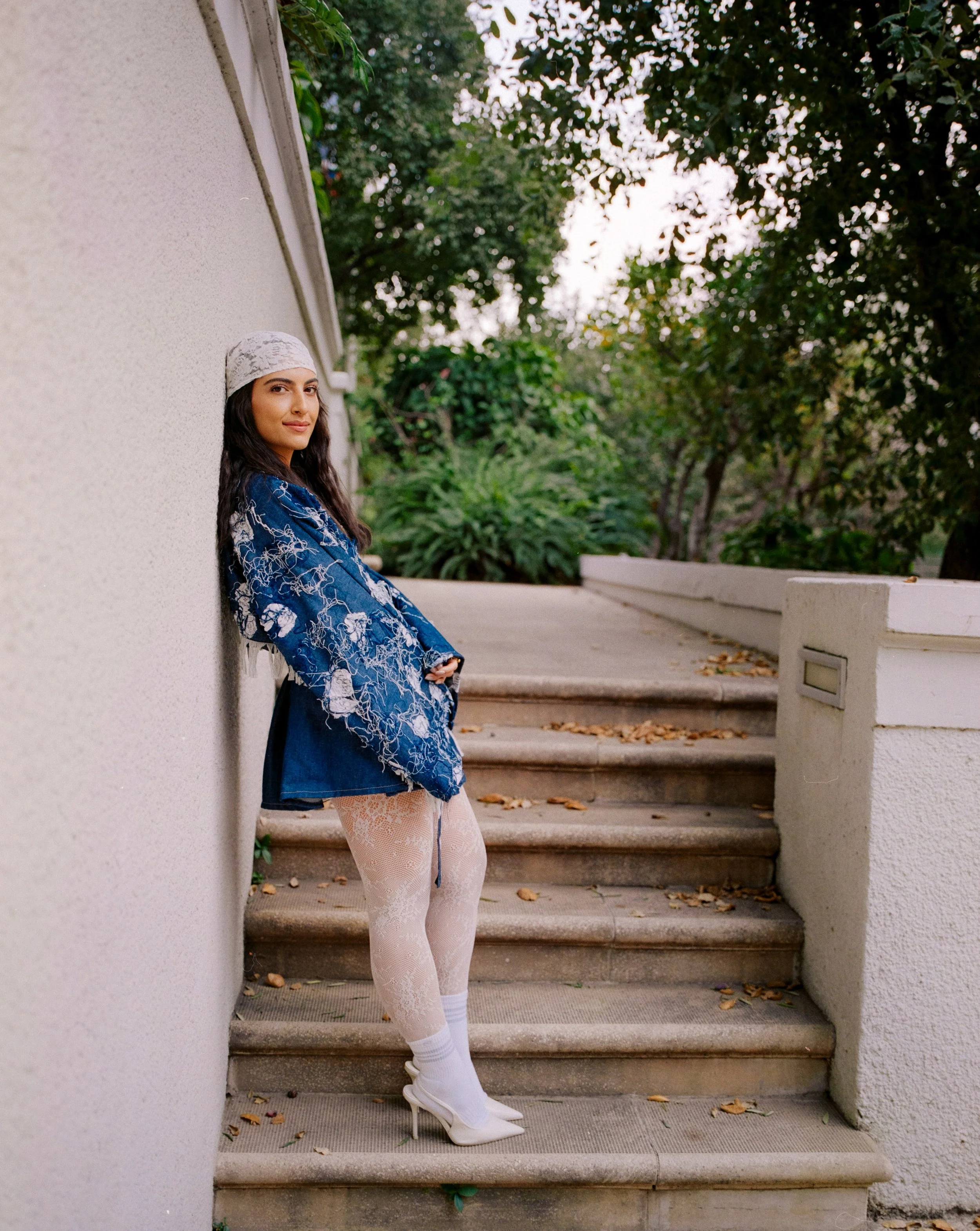 Young woman stands on outdoor stairs leaning against a wall, wearing a blue patterned jacket, white lace tights, white heels, a gray beanie, and smiling at the camera. Green trees and shrubs are in the background.