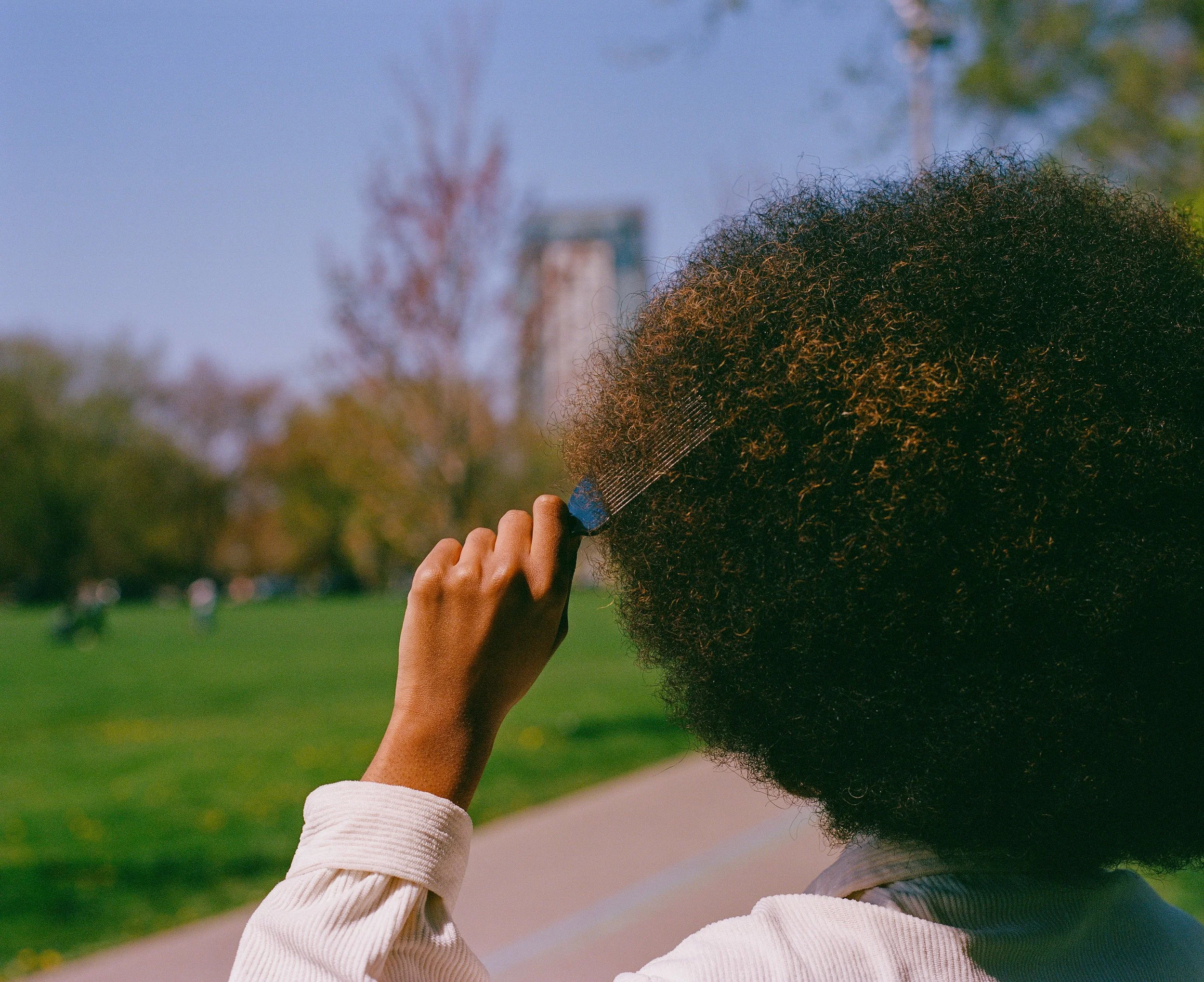Person with an afro hairstyle holding a small mirror or brush, in a park with trees and a city skyline in the background.