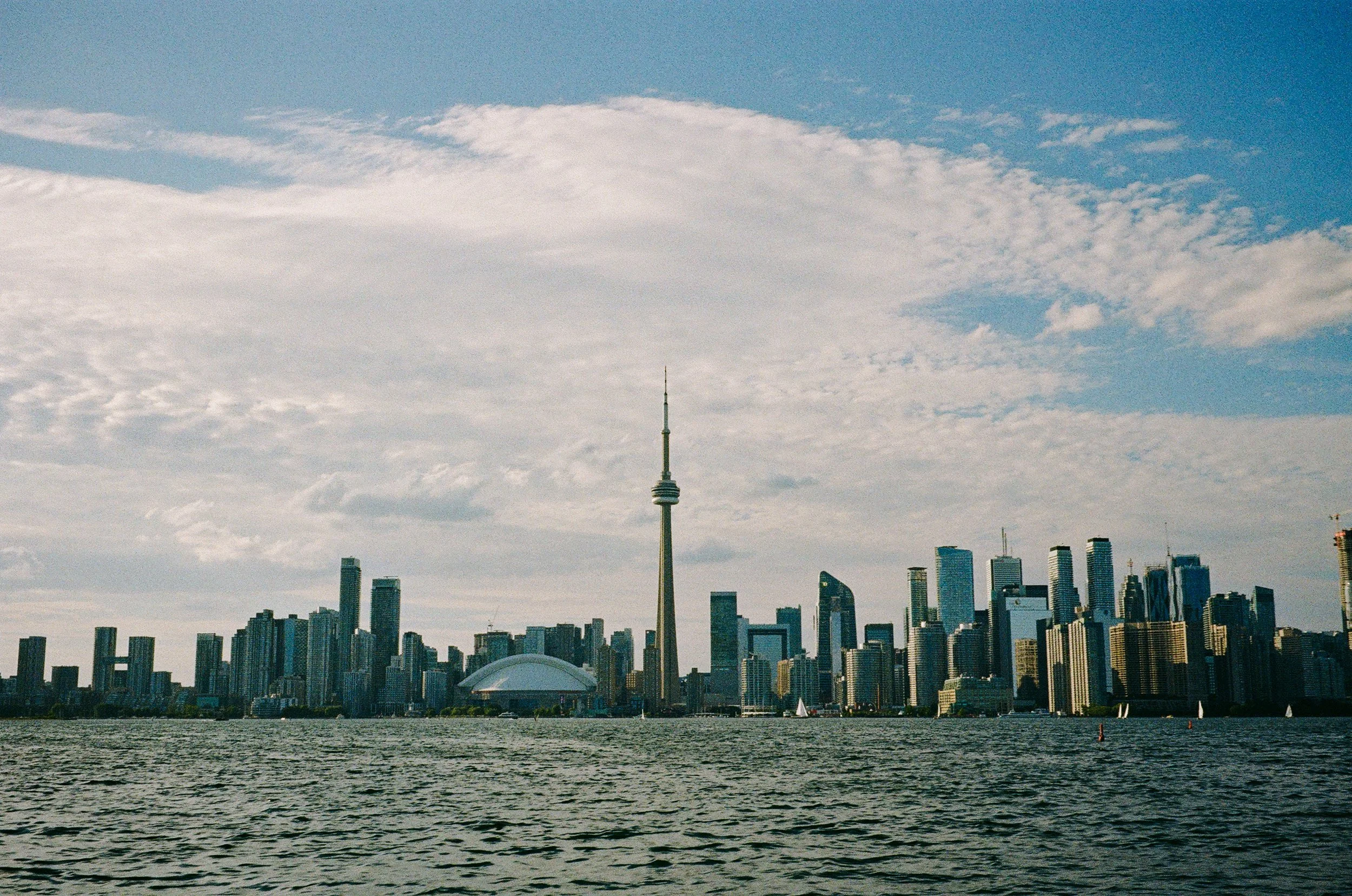 Skyline of downtown Toronto with CN Tower and Rogers Centre, viewed across water, under partly cloudy sky.