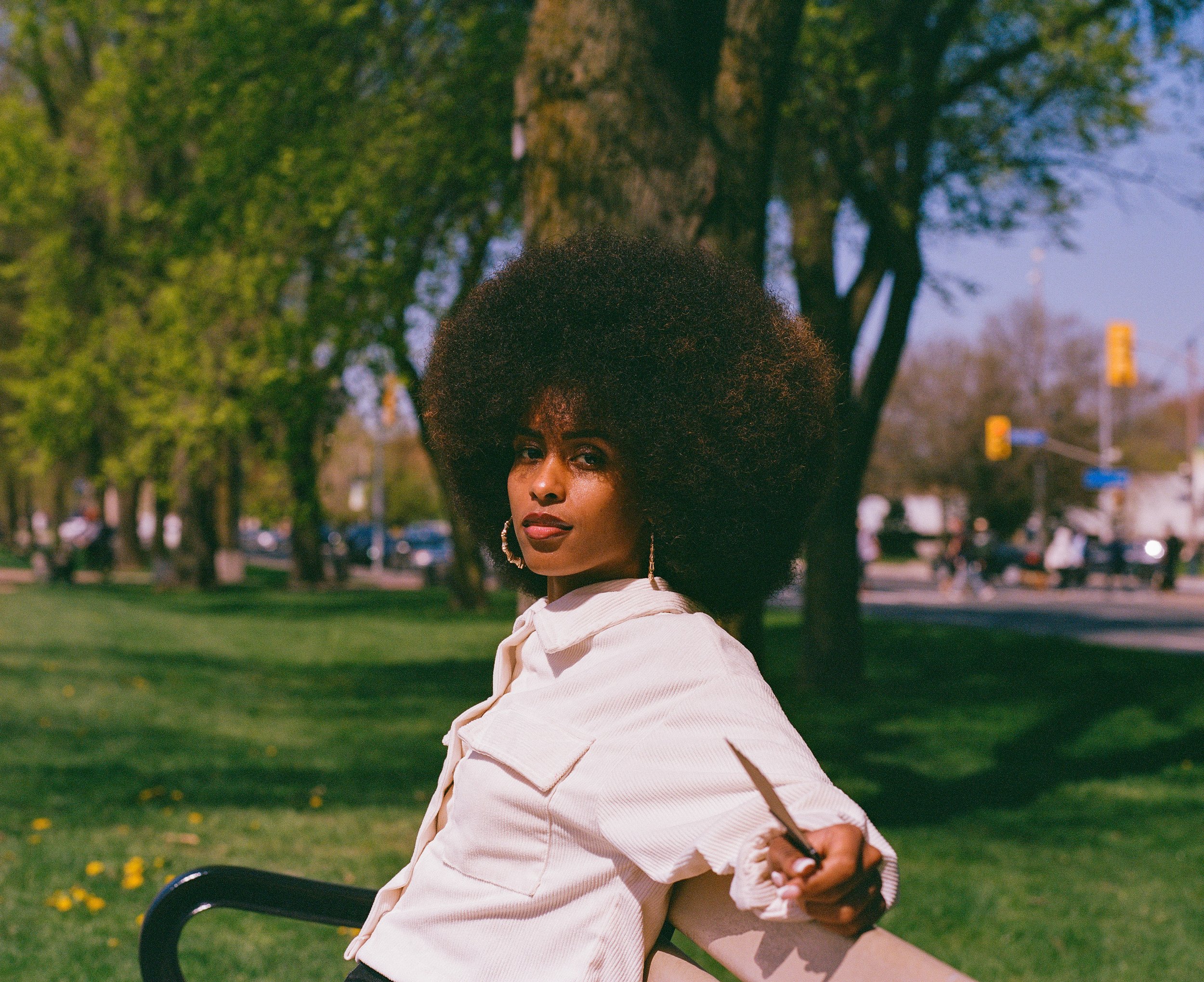 A woman with an Afro hairstyle and hoop earrings sitting on a park bench, holding a pen, with trees and a street with traffic lights in the background.