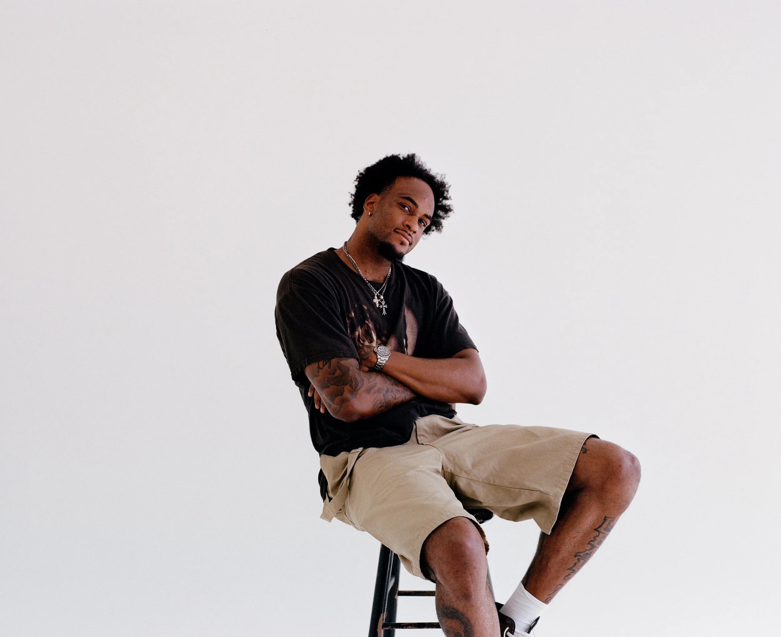 A young man with tattoos, wearing a black T-shirt and beige shorts, sitting on a stool with a white background, crossing his arms, looking at the camera.
