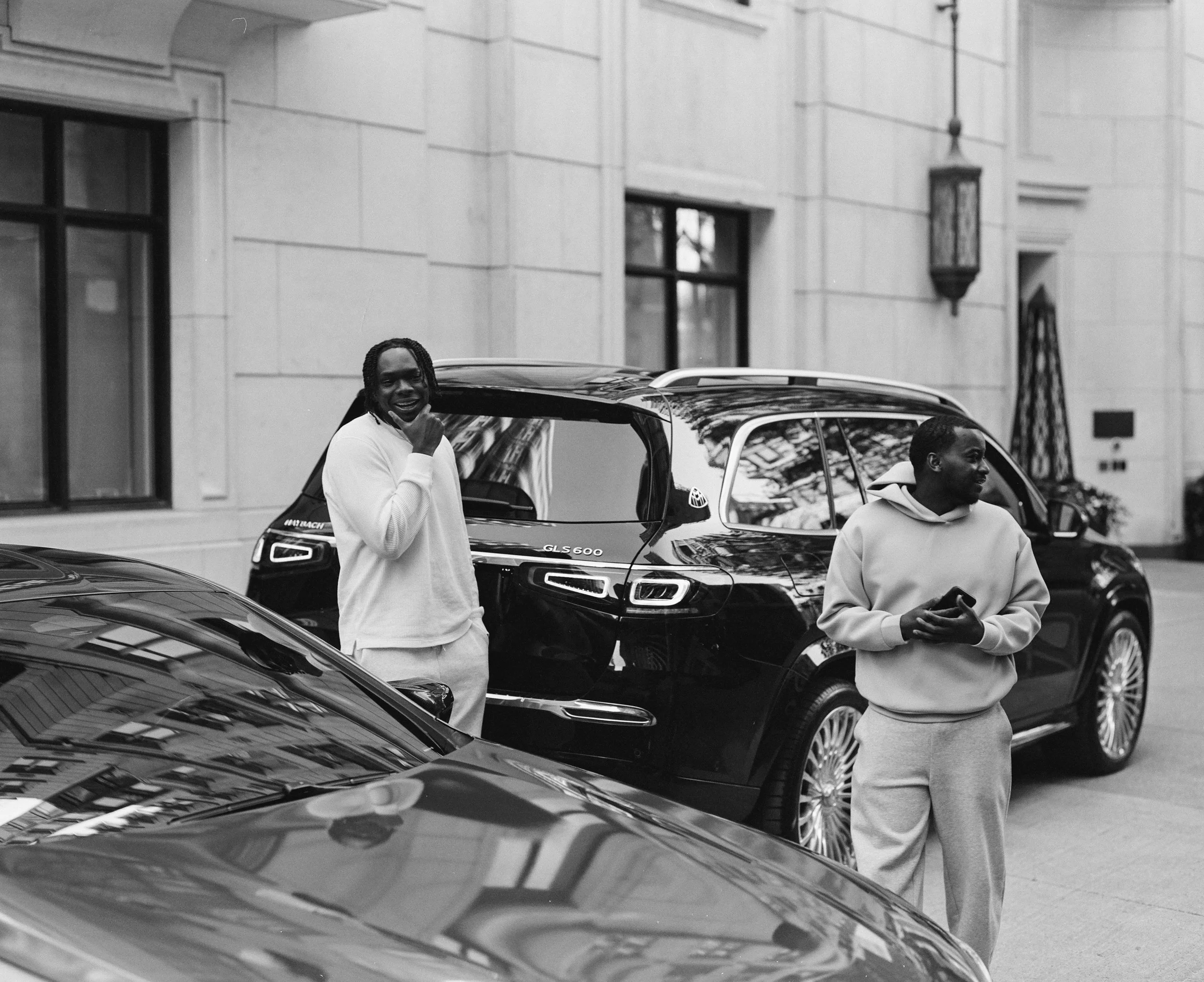 Two young men standing on a city street next to parked cars, one smiling and talking, the other holding a phone, with a large building with windows behind them.