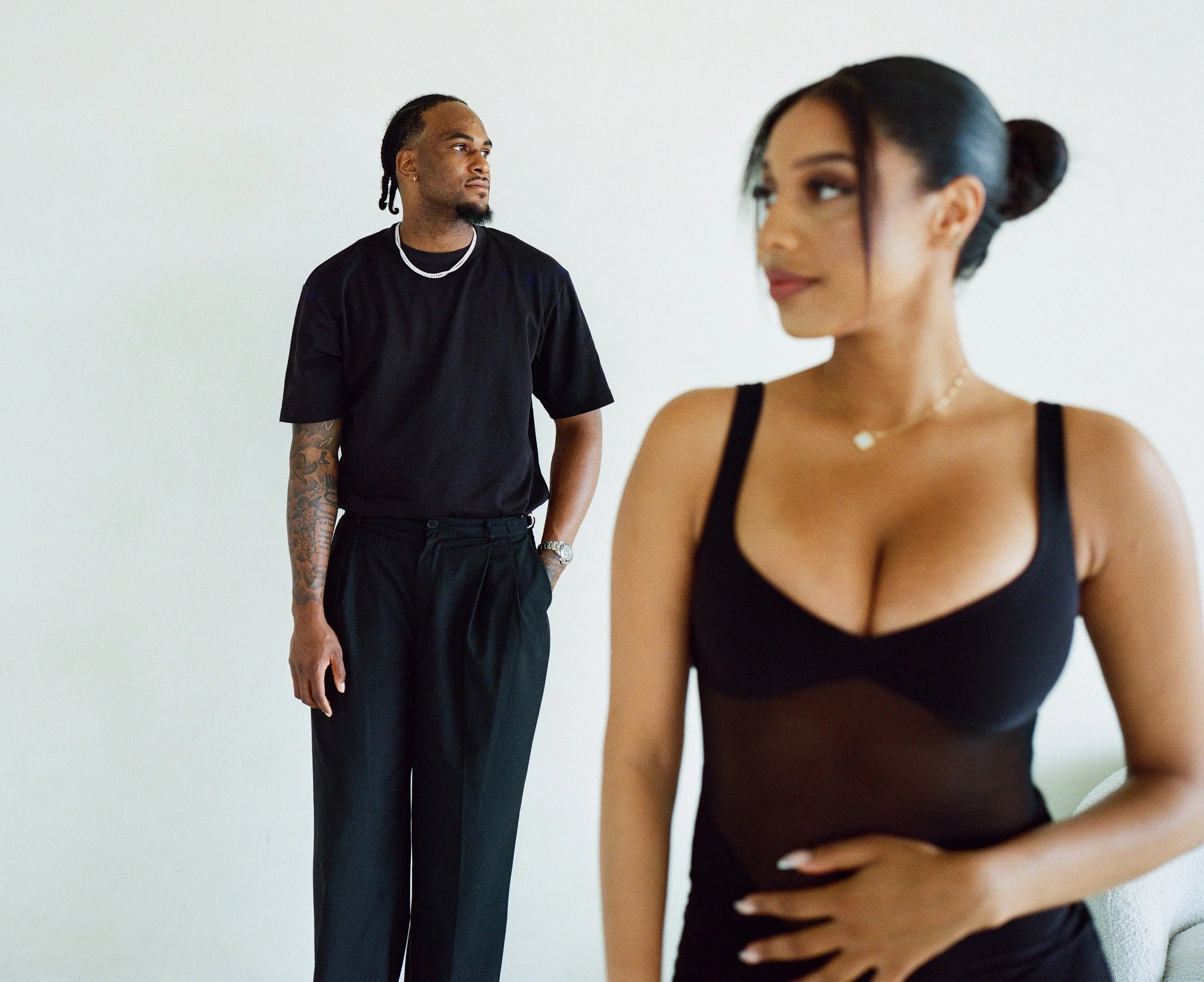 A man stands in the background looking to his left, wearing a black t-shirt, black pants, and silver jewelry, with tattoos on his left arm. A woman in the foreground, slightly out of focus, is wearing a black tank top and gazes to her right, with her