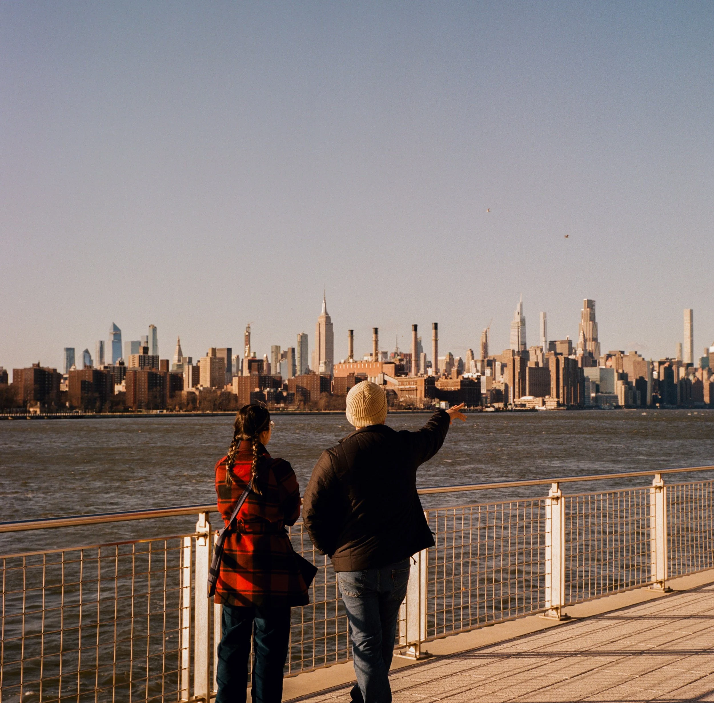 Two people stand by a waterfront railing, looking across a river at a city skyline with tall skyscrapers, including the Empire State Building. One person points towards the city.