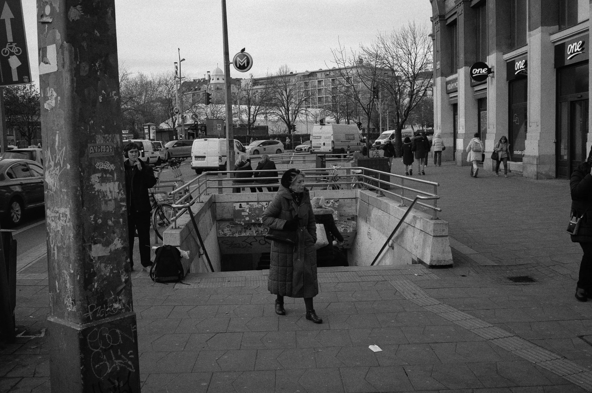 A street scene in black and white featuring pedestrians, cars, and a subway entrance with a woman standing near the stairs.
