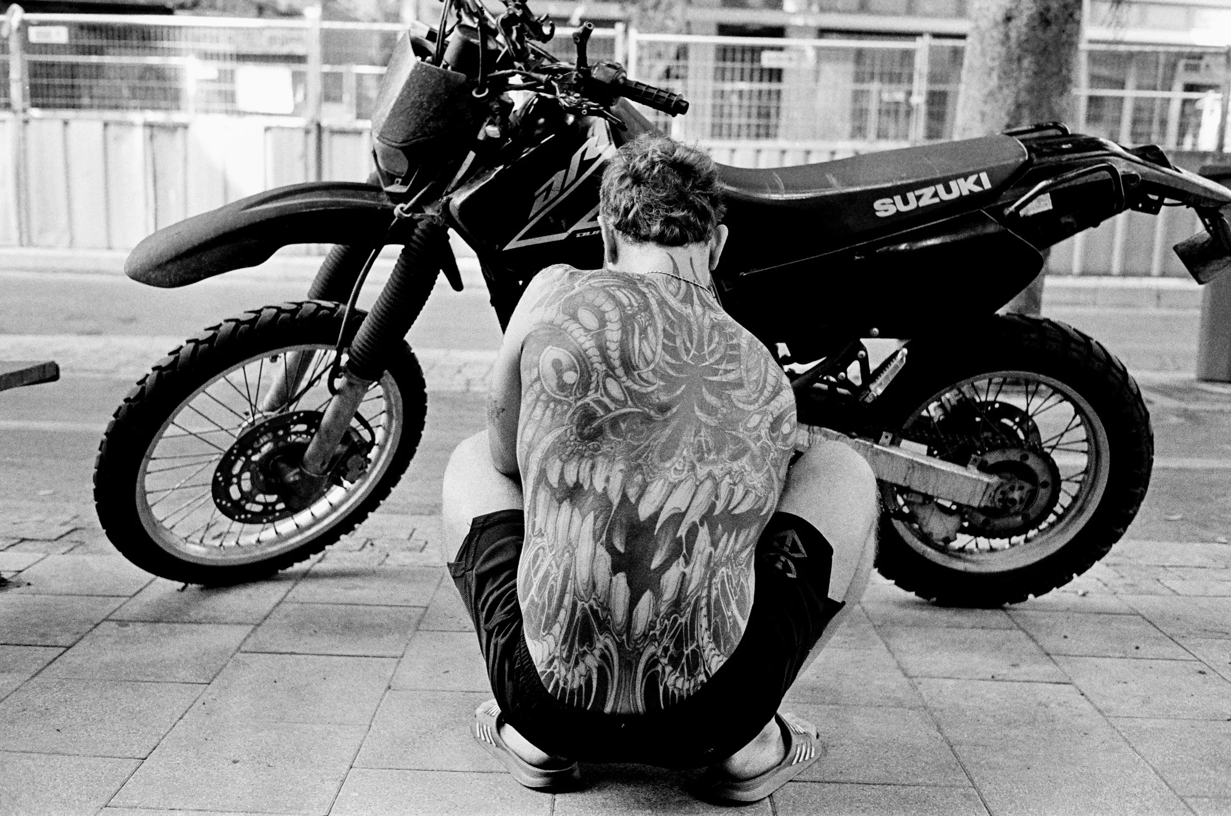 A person with tattoos on their back squatting in front of a Suzuki off-road motorcycle, on a city sidewalk with a railing in the background.