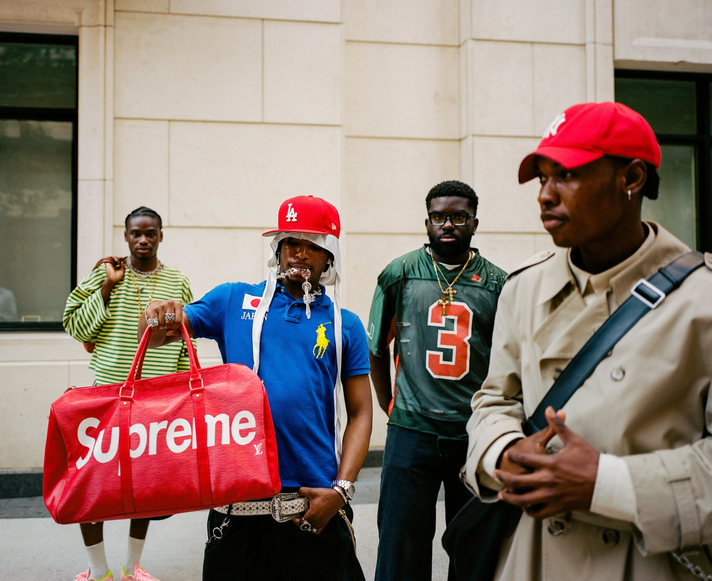 Group of four young men standing outside a building, one holding a large red Louis Vuitton Supreme duffel bag, dressed casually in streetwear and sports jerseys.