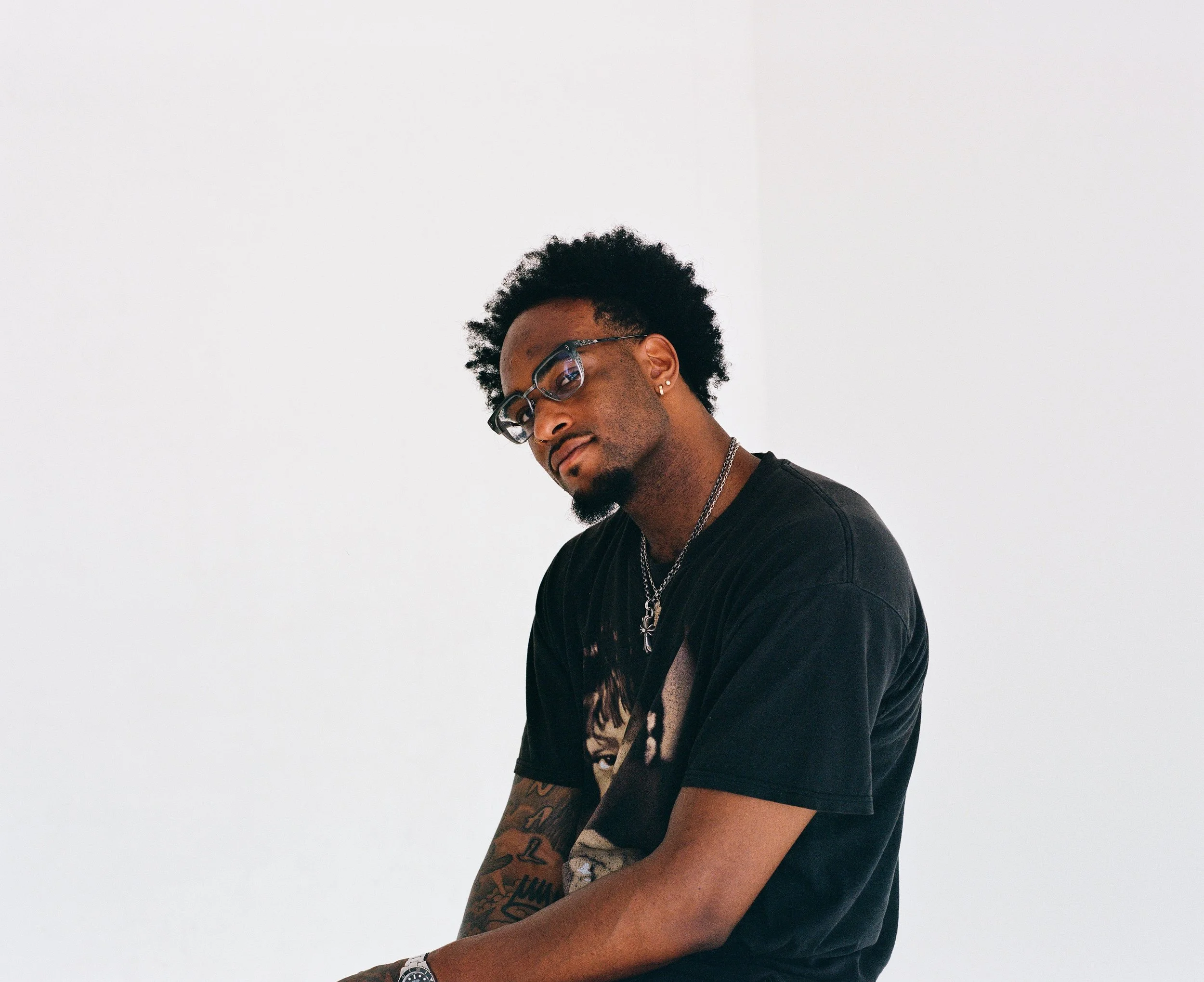 A young man with glasses and dark curly hair poses against a plain white background, wearing a black graphic t-shirt, multiple necklaces, and earrings.
