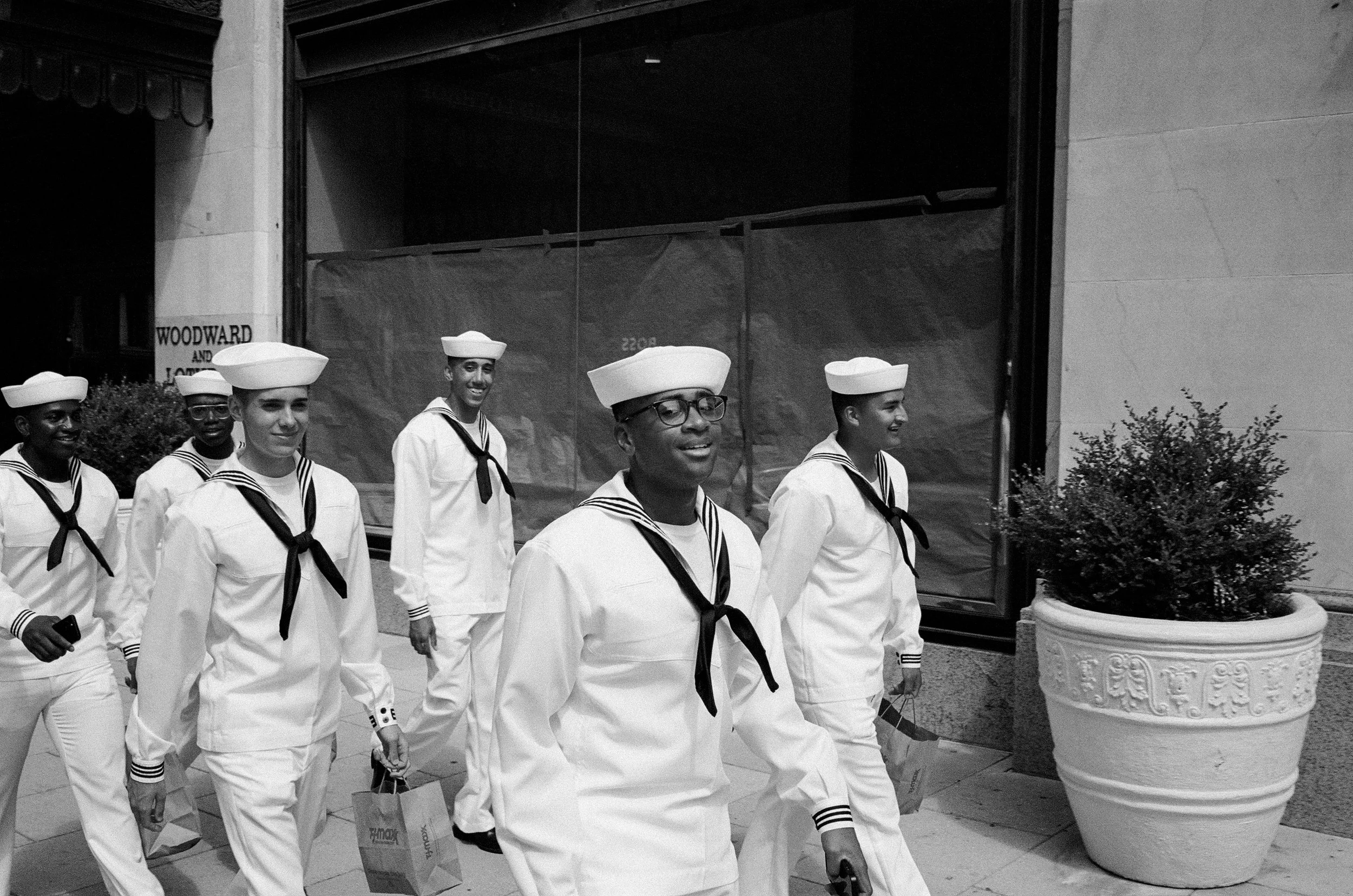 Group of people dressed in navy sailor uniforms walking outdoors in an urban area, carrying shopping bags.