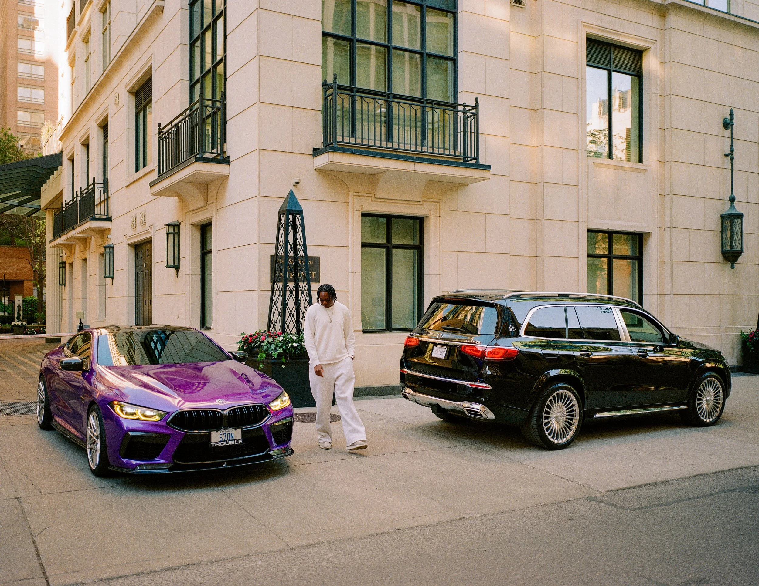 A man dressed in white standing between a purple sports car and a black SUV parked outside a beige apartment building, with flower pots and decorative lanterns on the sidewalk.