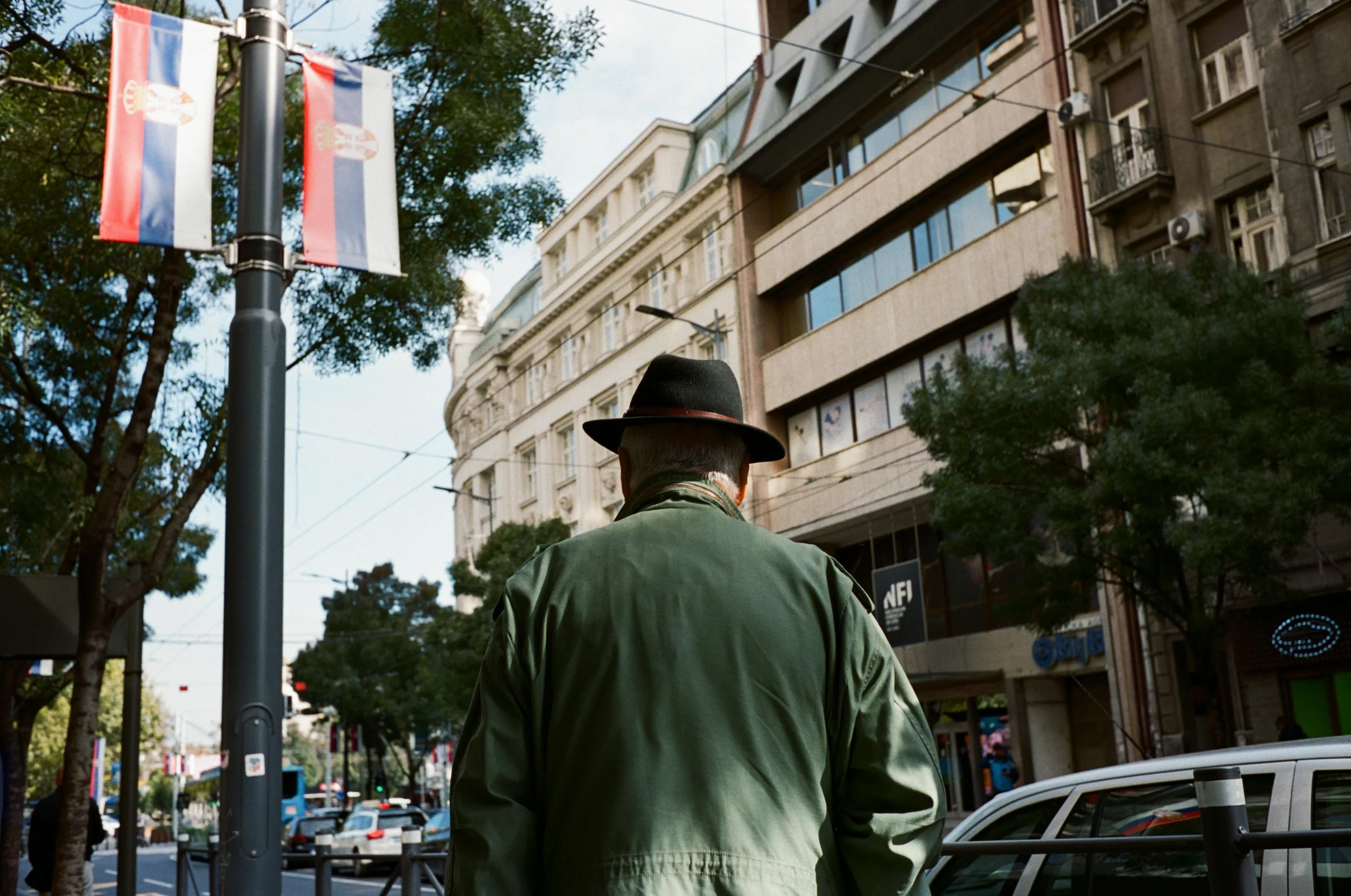 A man wearing a black hat and green jacket is standing on a city street with buildings, trees, cars, and flags in the background.