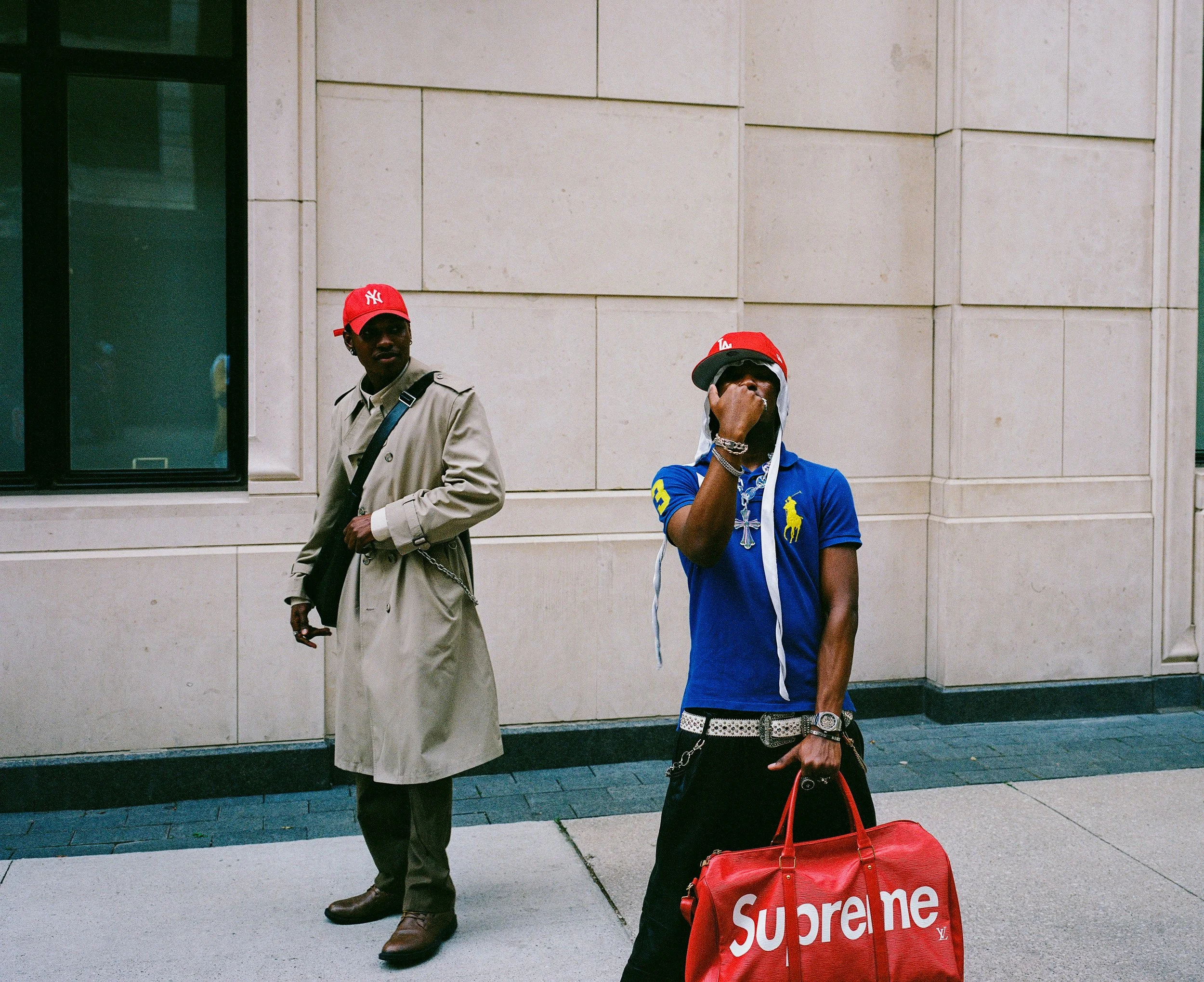Two men standing on a city sidewalk in front of a beige building. The man on the left wears a beige trench coat, dark pants, brown shoes, and a red baseball cap with the New York Yankees logo. The man on the right wears a blue polo shirt with yellow 