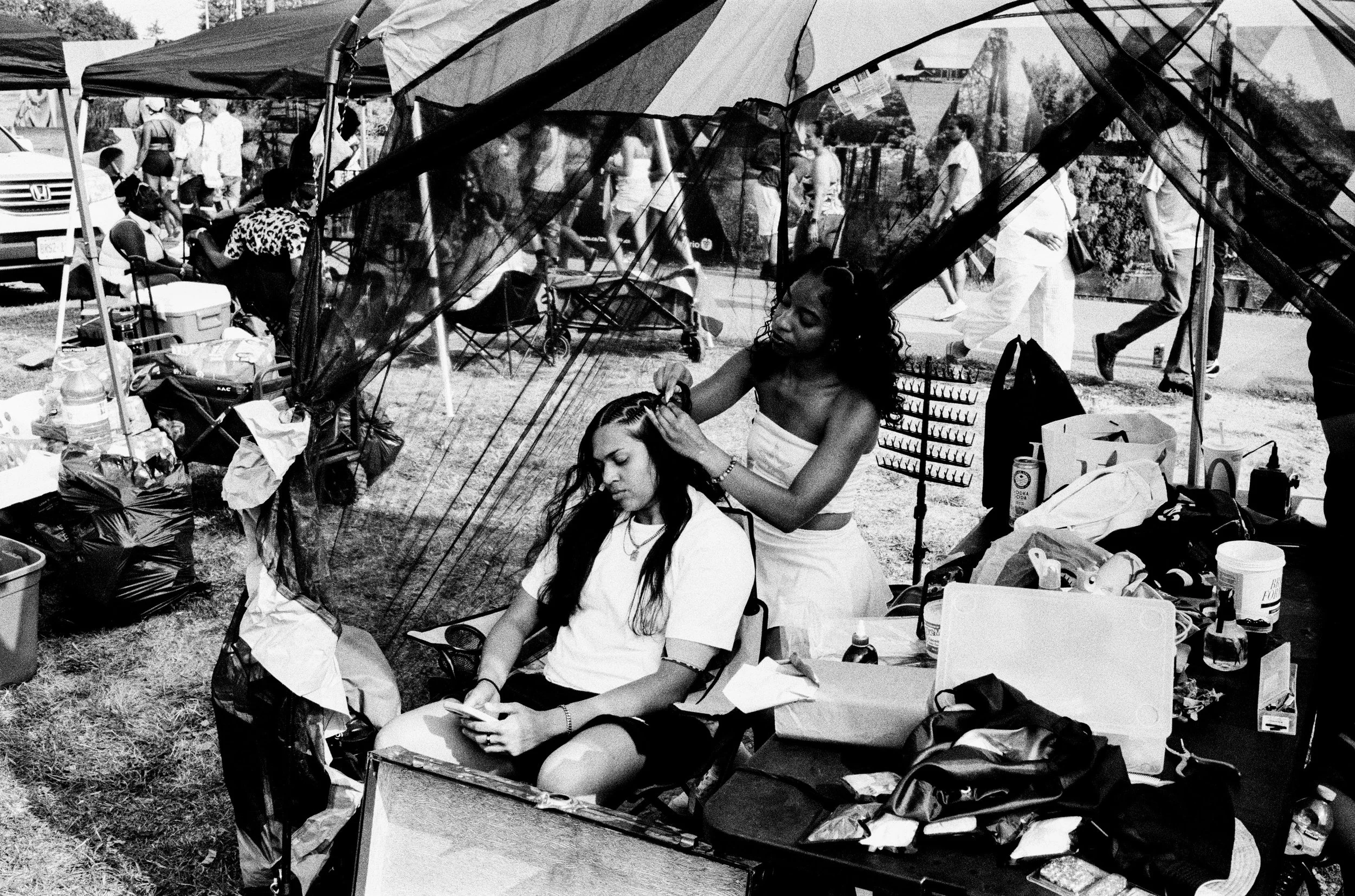 A woman with curly hair getting her hair styled by a hairdresser at an outdoor event. The hairdresser is standing and working on the woman's hair, who is seated and looking at her phone. There are tents and other people in the background, along with 
