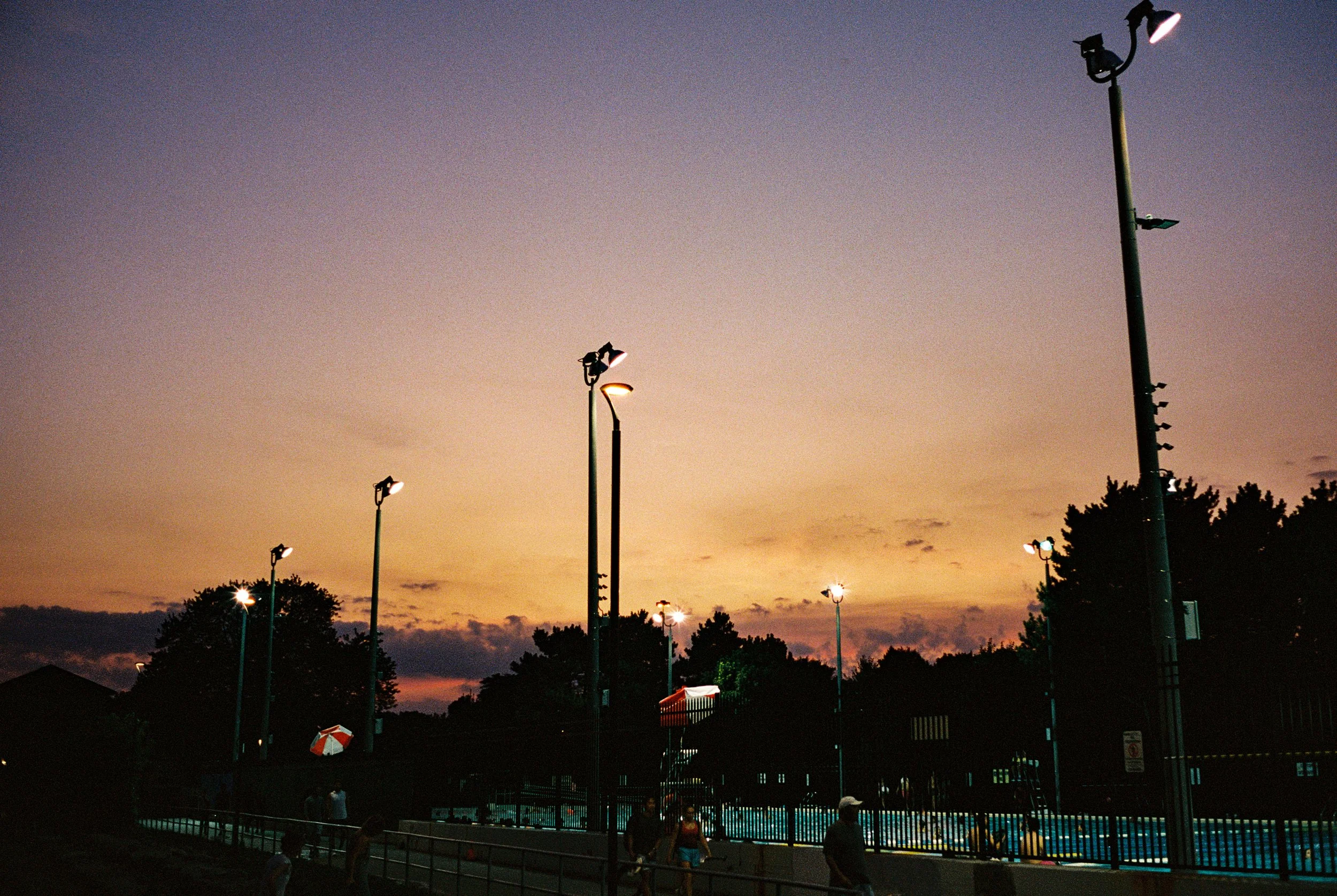 Outdoor swimming pool area at dusk with tall streetlights, trees, and a few people near the pool.