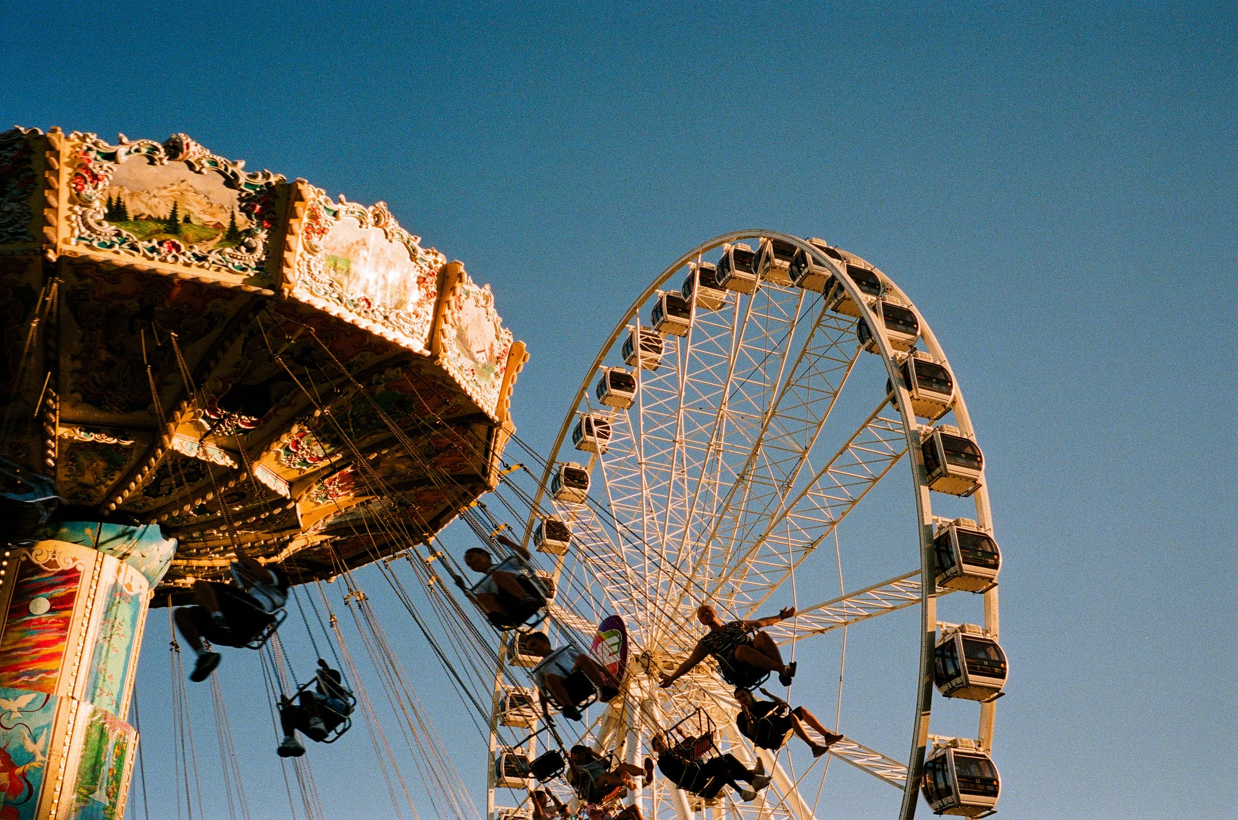 A colorful amusement park ride featuring a carousel and a Ferris wheel against a clear blue sky, with people enjoying the rides.
