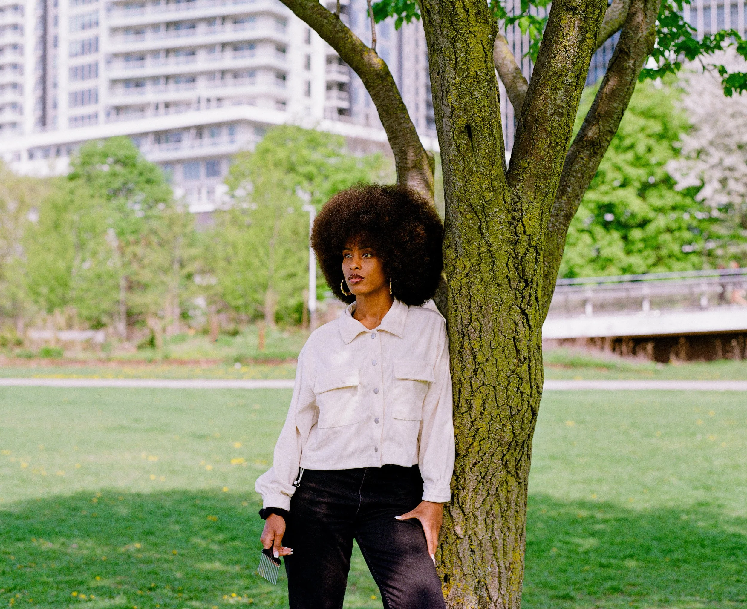 A woman with large curly hair leaning against a tree in a park with green grass and trees, and tall modern buildings in the background.