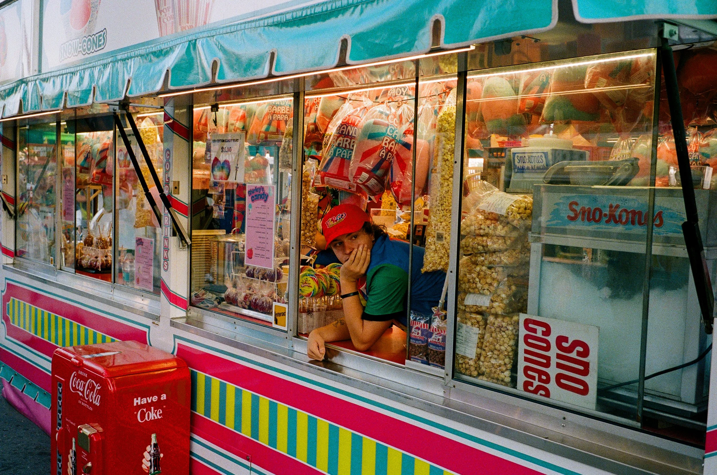 A girl leaning on a snack stand counter with her hand supporting her face, surrounded by popcorn and candy, wearing a red cap and a green shirt.