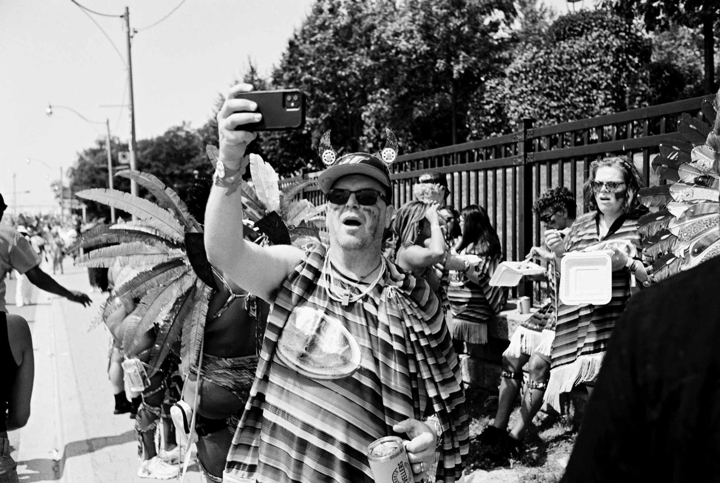 People at a parade, some dressed in costumes with feathers, one person taking a selfie, others holding food and drinks, some wearing sunglasses, behind a black fence and trees.