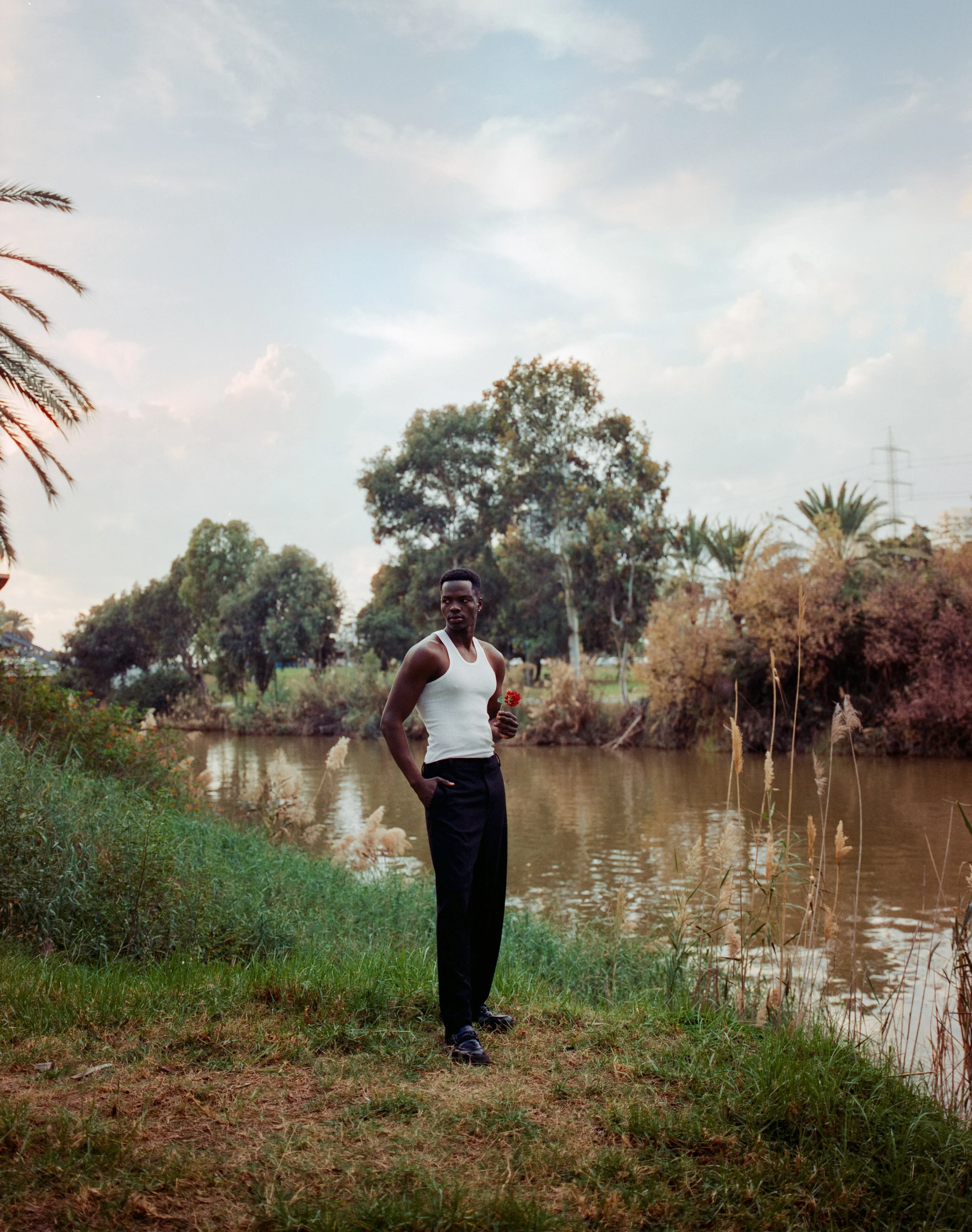 A man in a white tank top and dark pants standing by a river, holding a flower, with trees and a partly cloudy sky in the background.