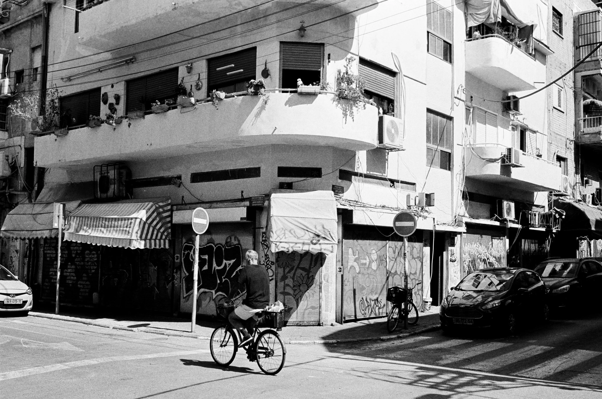 Black and white photo of a man riding a bicycle on a city street. The building in the background has multiple balconies with plants, air conditioning units, and graffiti-covered metal shutters. Multiple parked cars are along the street, and two 'no e