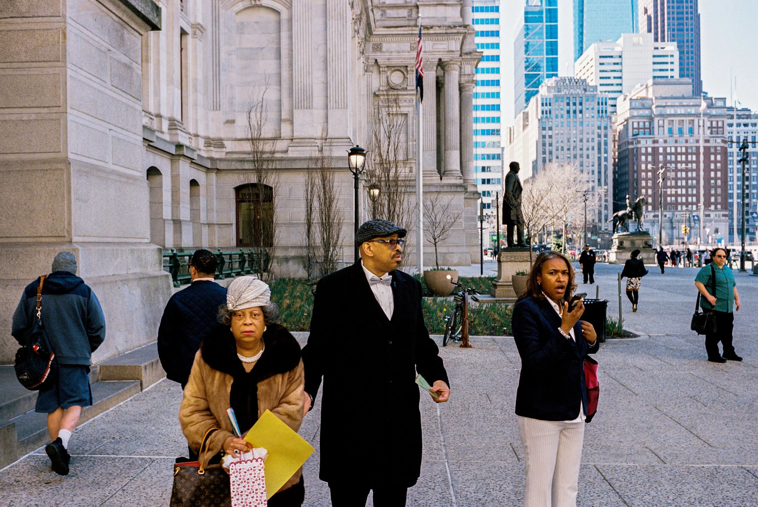 People standing on a city sidewalk in front of a historic building with statues and tall modern skyscrapers in the background.