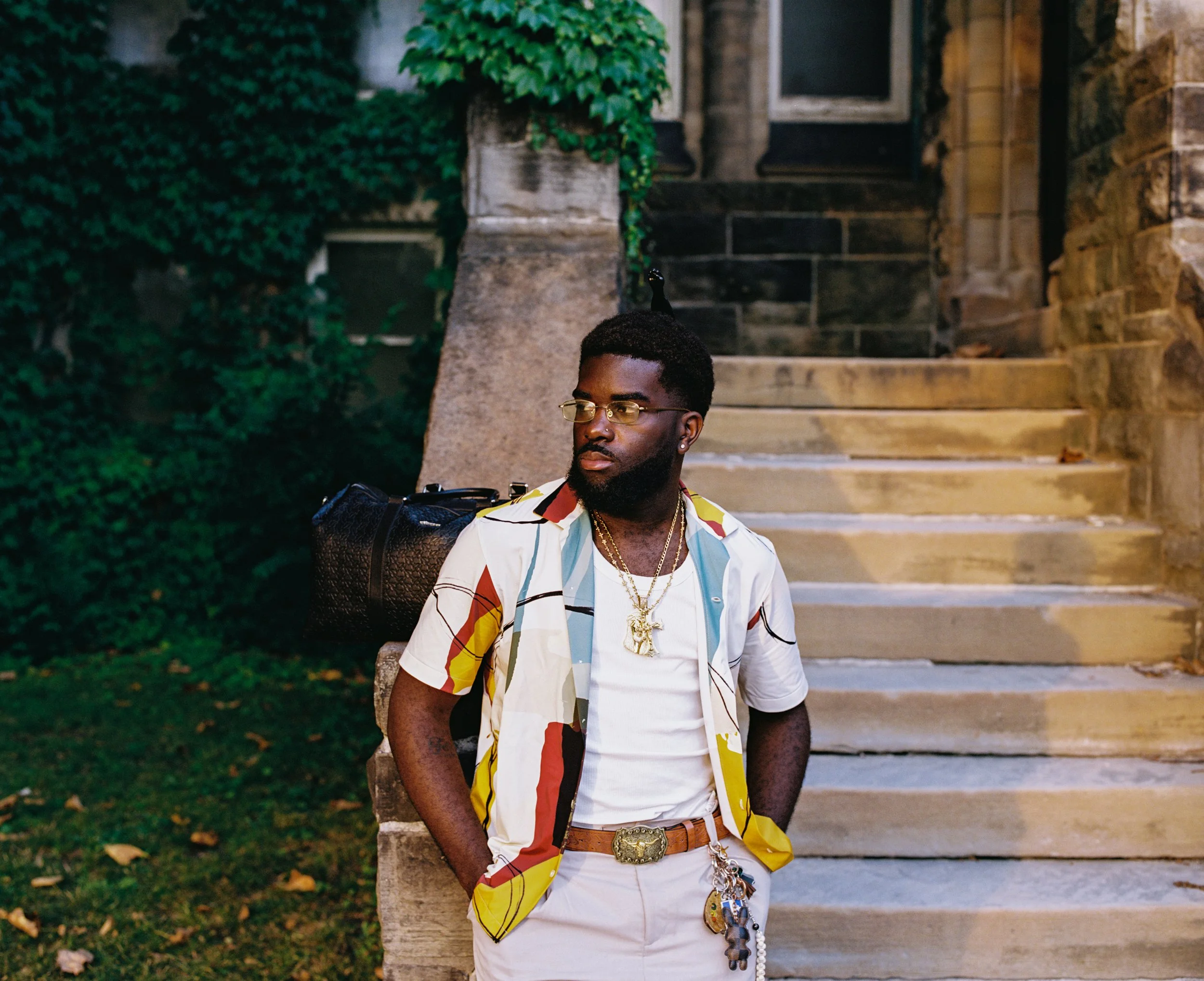 A man with dark skin, wearing a white t-shirt, colorful patterned short-sleeve shirt, sunglasses, and gold jewelry, including chains and a belt, stands outdoors on a stone staircase with green foliage and an old building in the background.